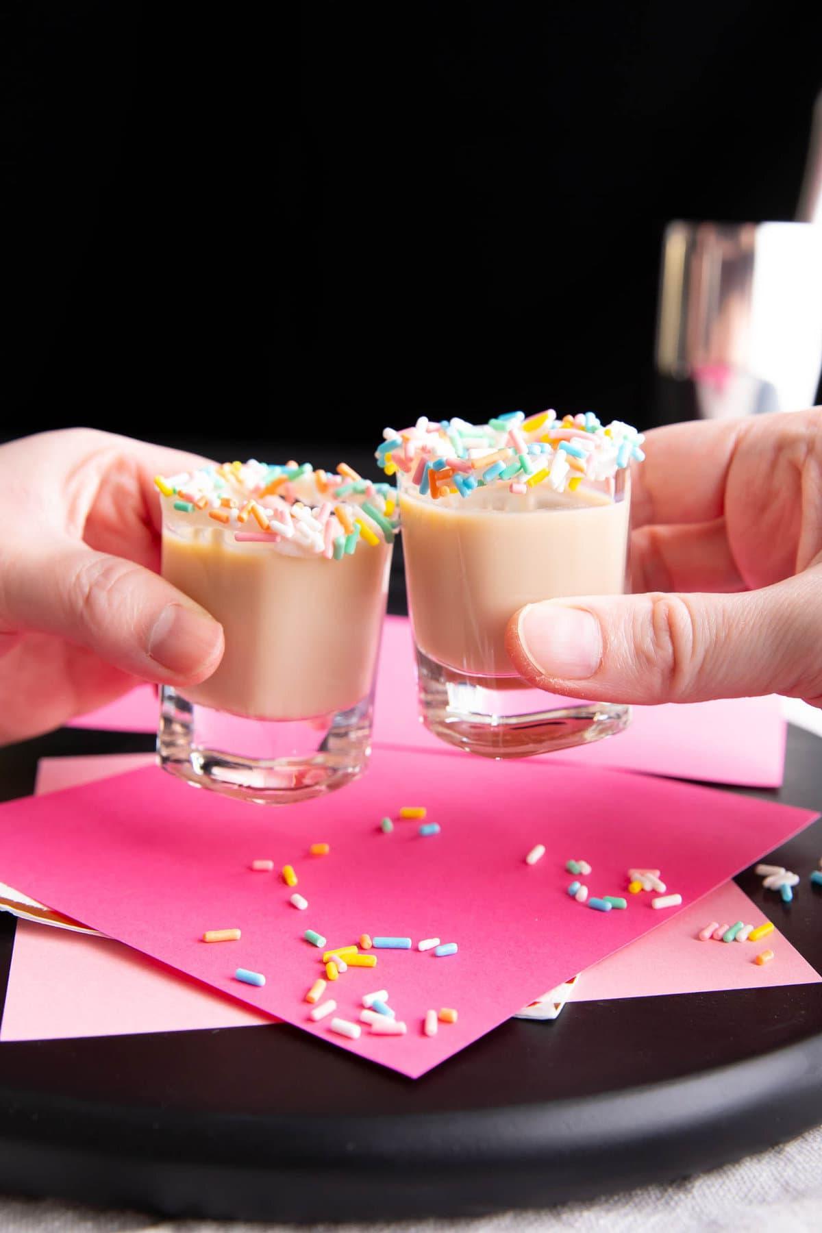 Geburtstagskuchen-Shot-Rezept 2 Zwei Hände halten Geburtstagskuchen-Shots, stoßen Schnapsgläser an