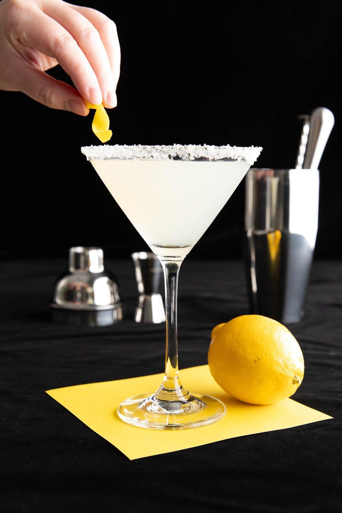 Das beste Lemon Drop Martini Rezept 3 Hand platziert eine Zitronenzeste auf dem Zitronen-Zucker-Rand eines Lemon Drops.