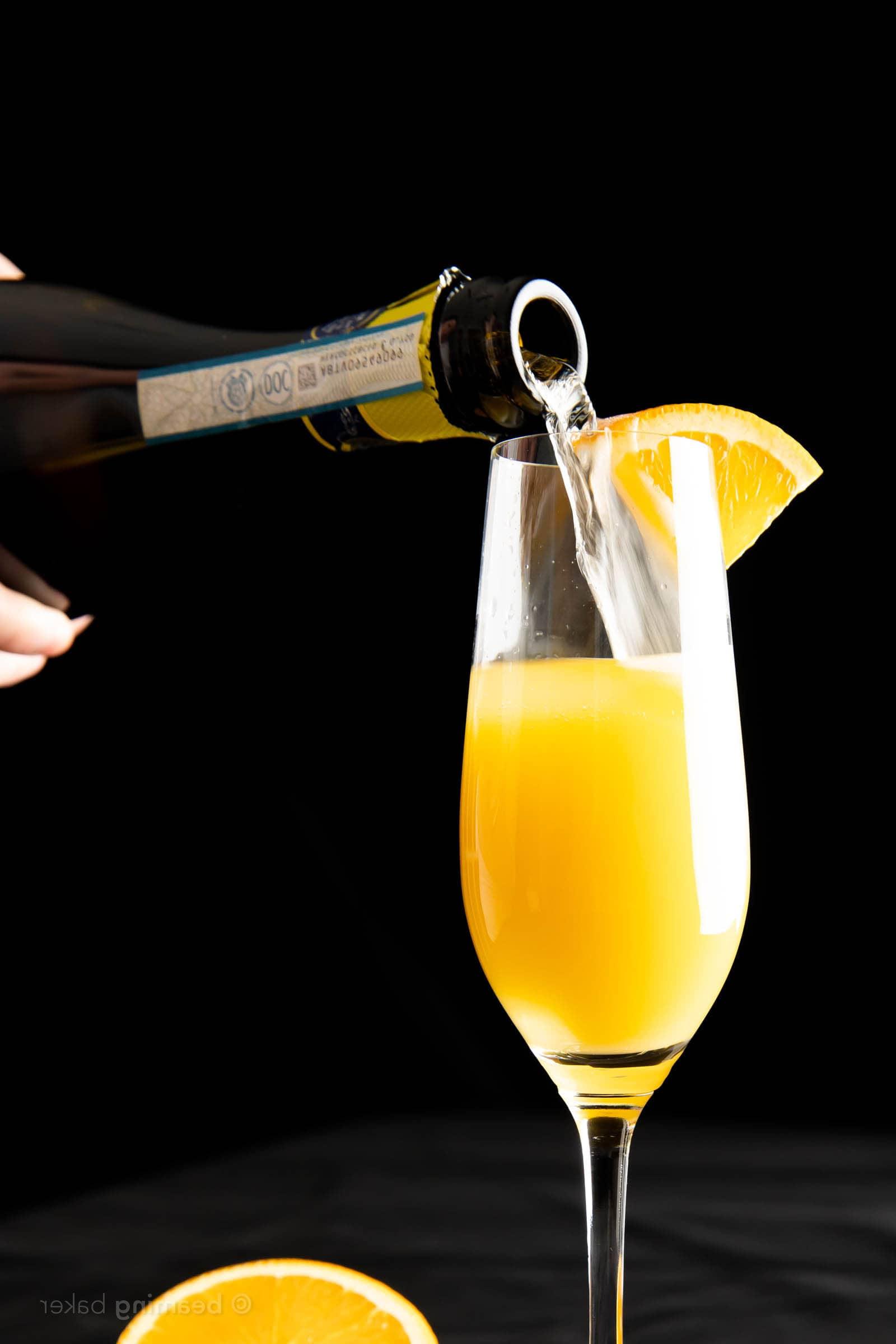 Nahaufnahme einer Hand, die Prosecco in ein halb mit Orangensaft gefülltes Sektglas für einen Mimosa gießt