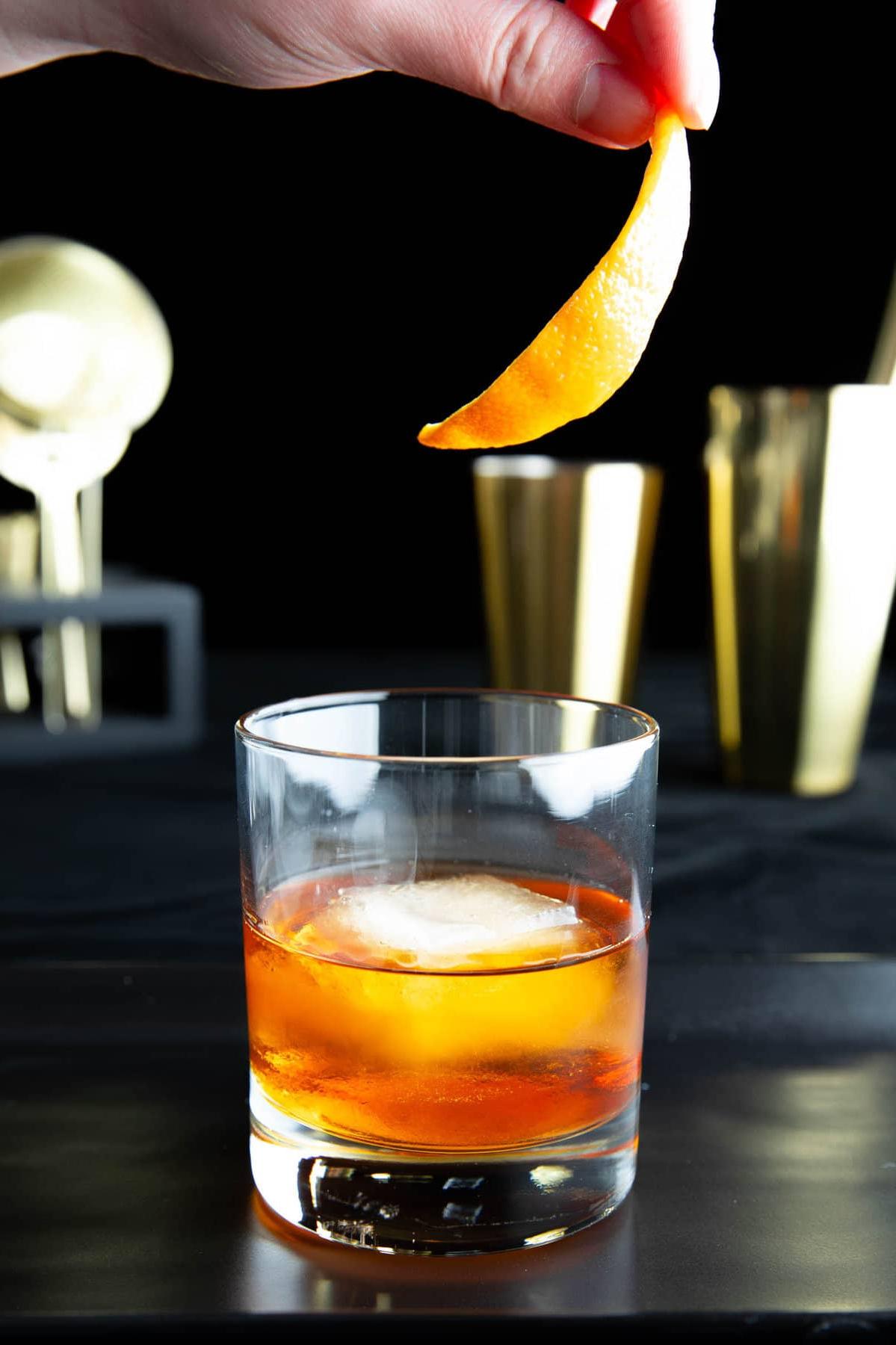 Old Fashioned Getränkerezept 3 Eine Hand, die eine Orangenschale als Garnitur zu einem Old Fashioned Cocktail hinzufügt