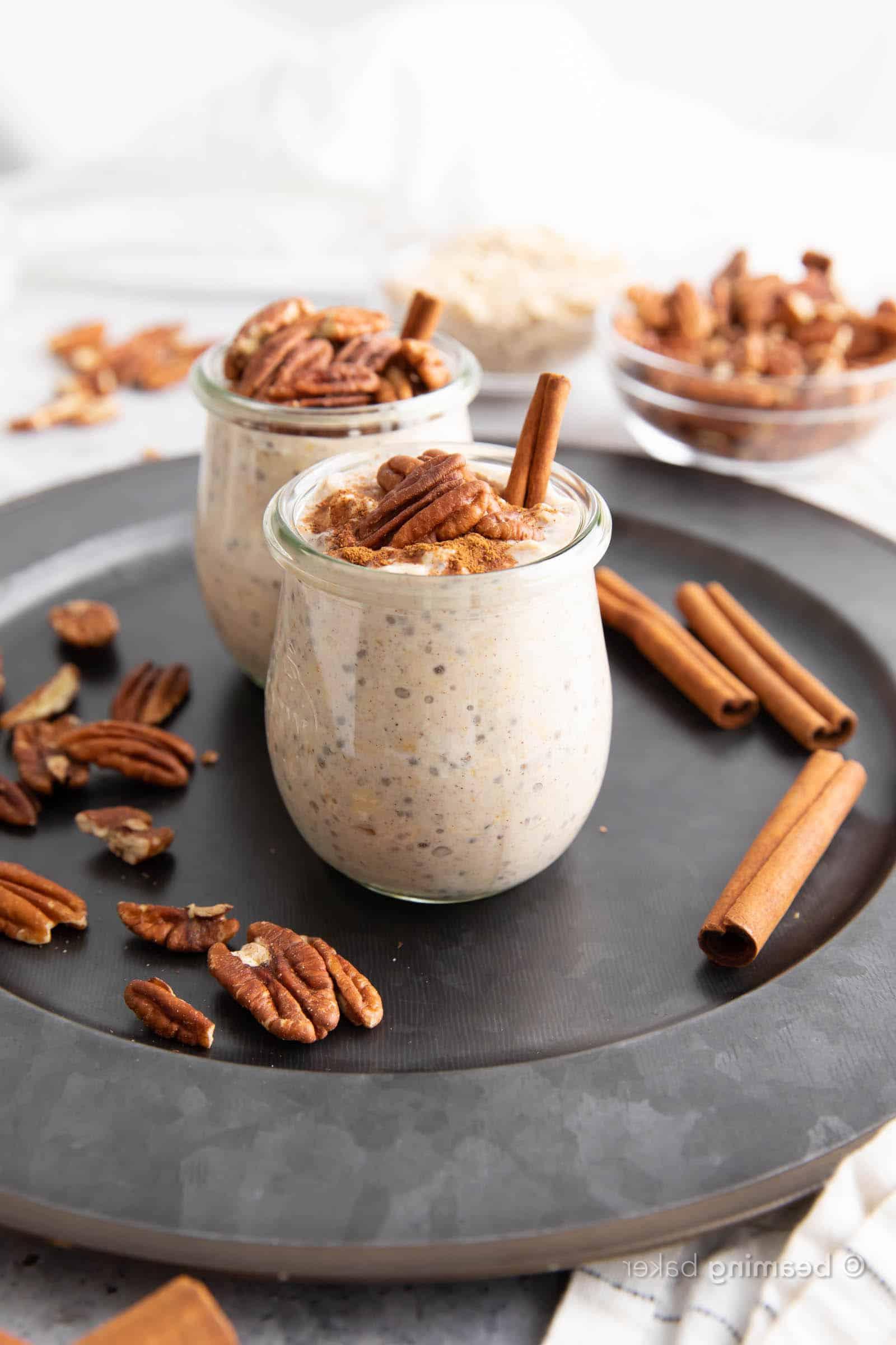 Ahorn-Pekannuss-Overnight-Oats 4 Zwei Gläser Ahorn-Pekannuss Overnight Haferflocken, serviert auf einem Servierteller mit Zimtstangen und Pekannusshälften.