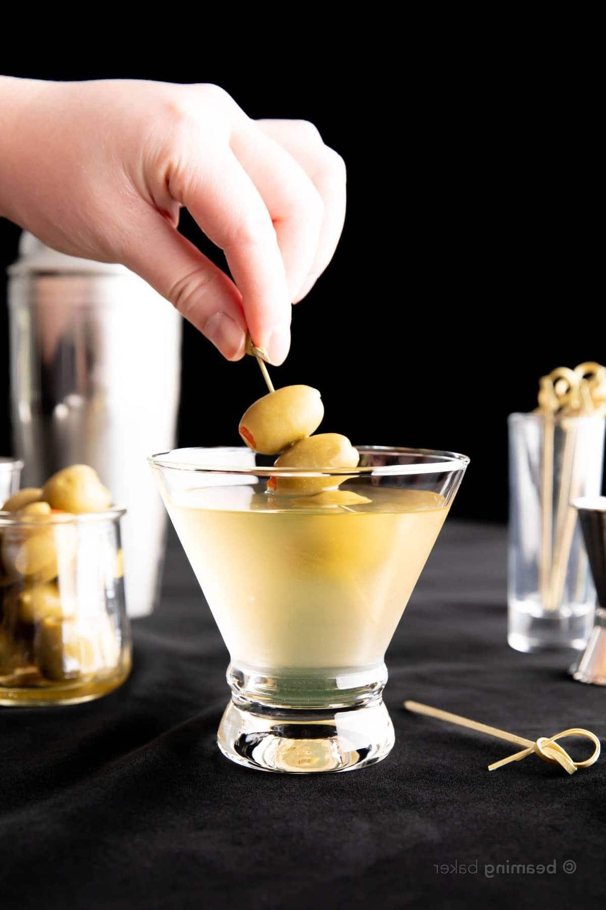 Extra Schmutziger Martini 7 Hand hält Oliven-Spieß, der fast im Cocktail versenkt ist