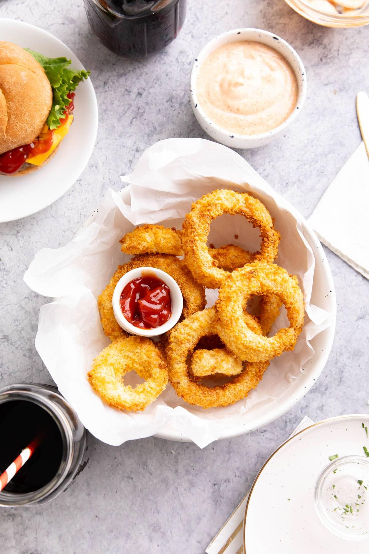 **Selbstgemachte Heißluftfritteusen-Zwiebelringe** 9 Ein Esstisch mit einer Schüssel knuspriger Zwiebelringe aus der Heißluftfritteuse, Zwiebelringsauce, Ketchup, einem Burger und Gläsern mit Limonade.