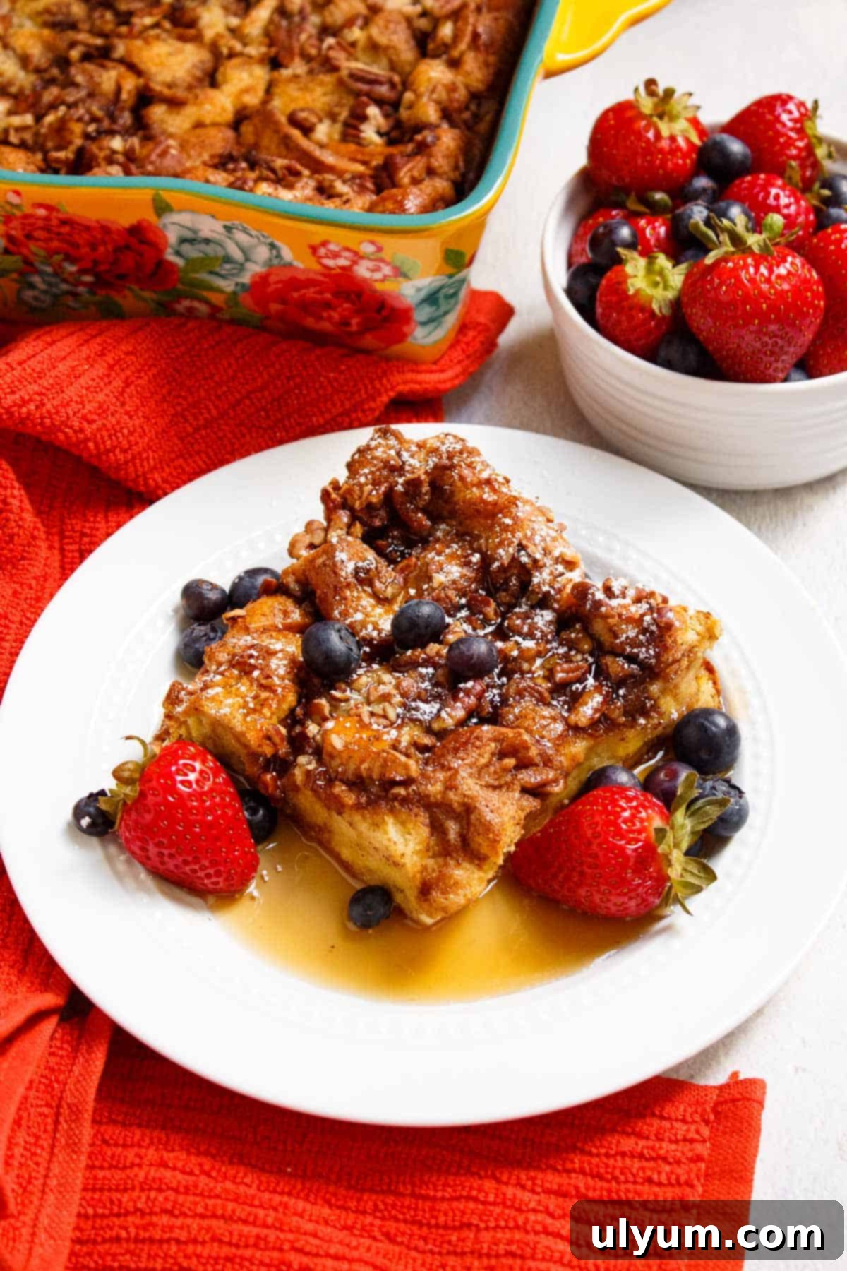 Square of French toast casserole served with berries, powdered sugar, and maple syrup.