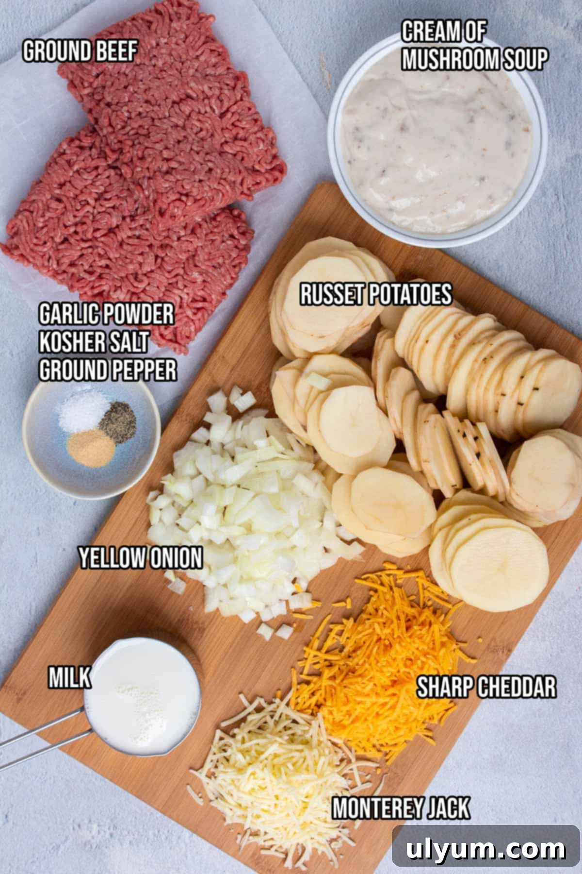 All the ingredients for Crockpot Hamburger Potato Casserole laid out on a kitchen counter: ground beef, onions, russet potatoes, cream of mushroom soup, milk, spices, and shredded cheeses.