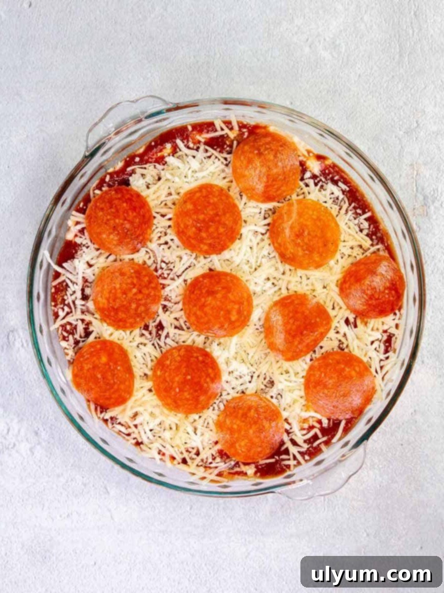 Top down view of pizza dip in a pie plate before it is baked, showing the layers of cheese and pepperoni slices.