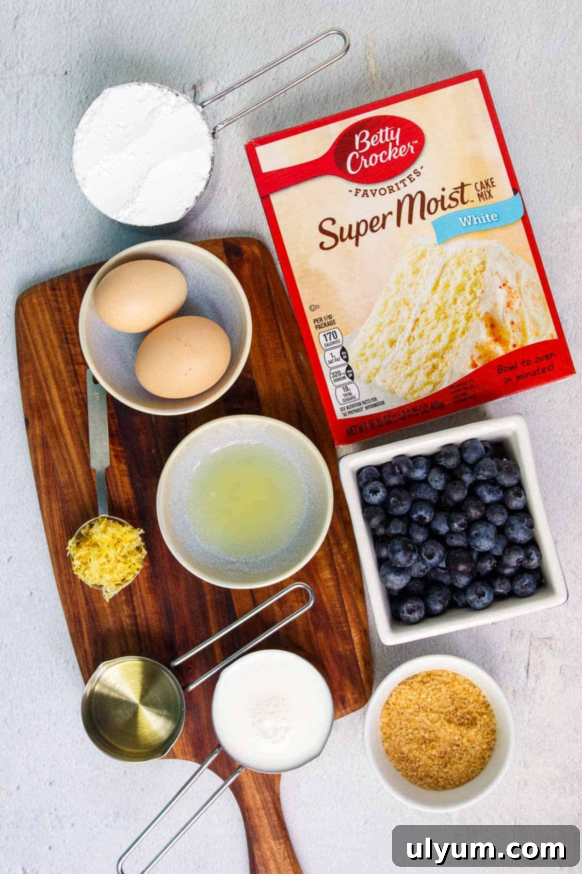 Various ingredients for cake mix blueberry muffins laid out on a table: a box of white cake mix, fresh blueberries, eggs, milk, vegetable oil, lemon, turbinado sugar, powdered sugar.