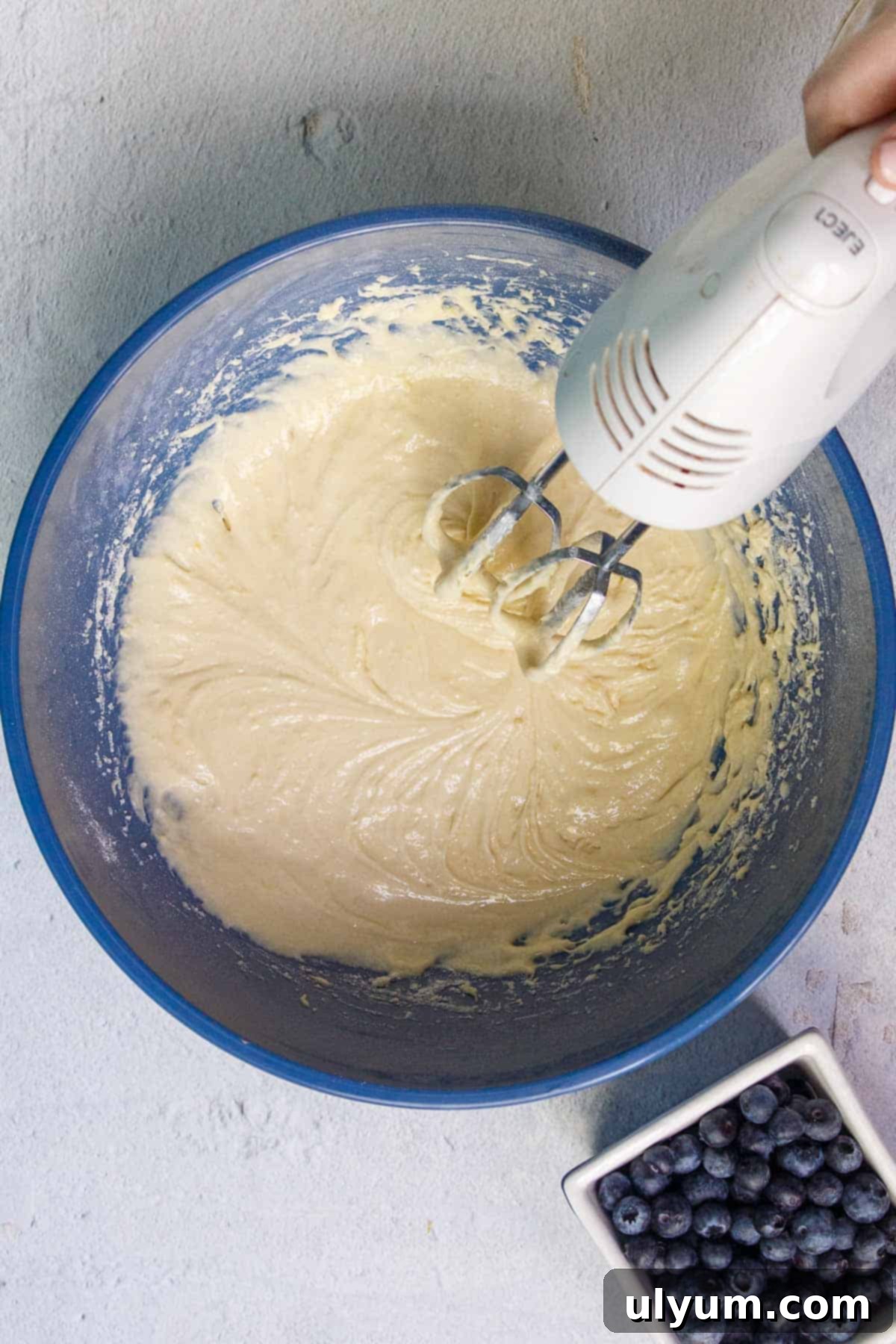 Beating cake mix blueberry muffin batter with a hand mixer in a large mixing bowl, showing the consistency of the wet batter.