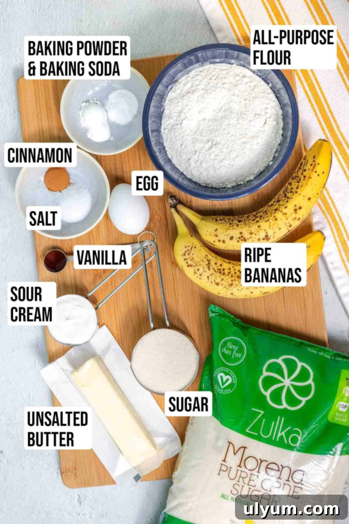 Individual ingredients for the banana coffee cake laid out on a table, including softened butter, sugars, mashed bananas, egg, sour cream, vanilla, flour, leavening agents, cinnamon, and salt.