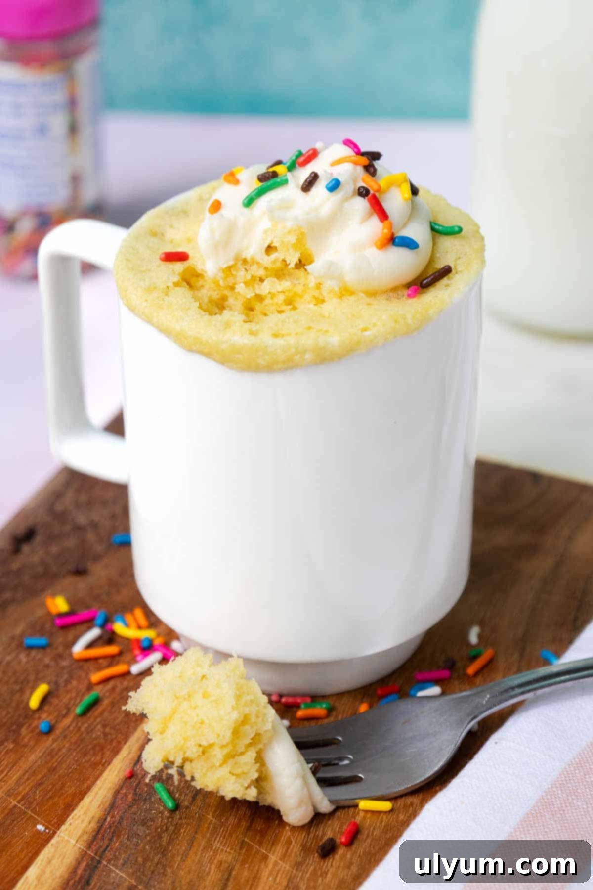 Yellow mug cake topped with buttercream frosting and sprinkles. A bite of cake rests on a fork to show the light texture.