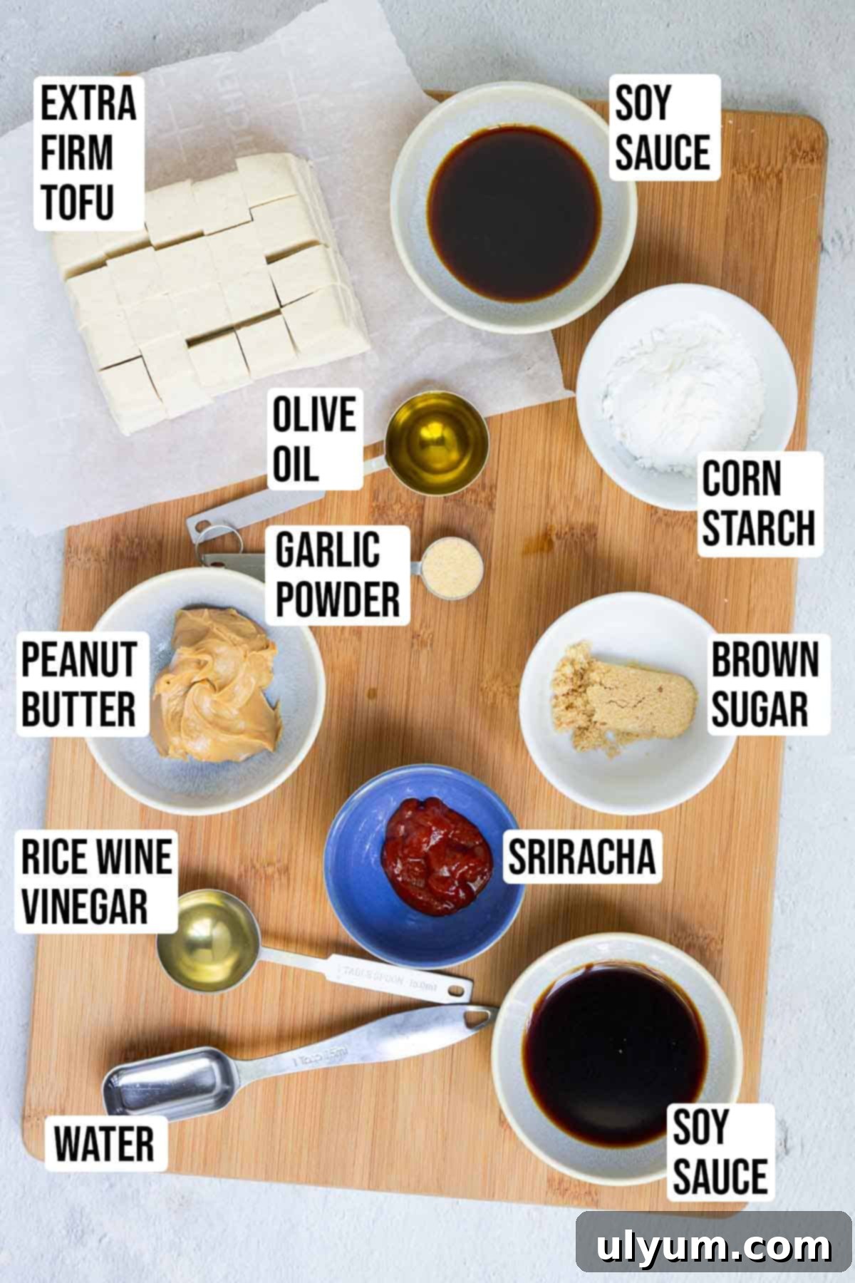 Ingredients for baked tofu with peanut sauce, including cubed tofu, soy sauce, olive oil, garlic powder, corn starch, peanut butter, rice vinegar, brown sugar, and sriracha laid out on a wooden board.