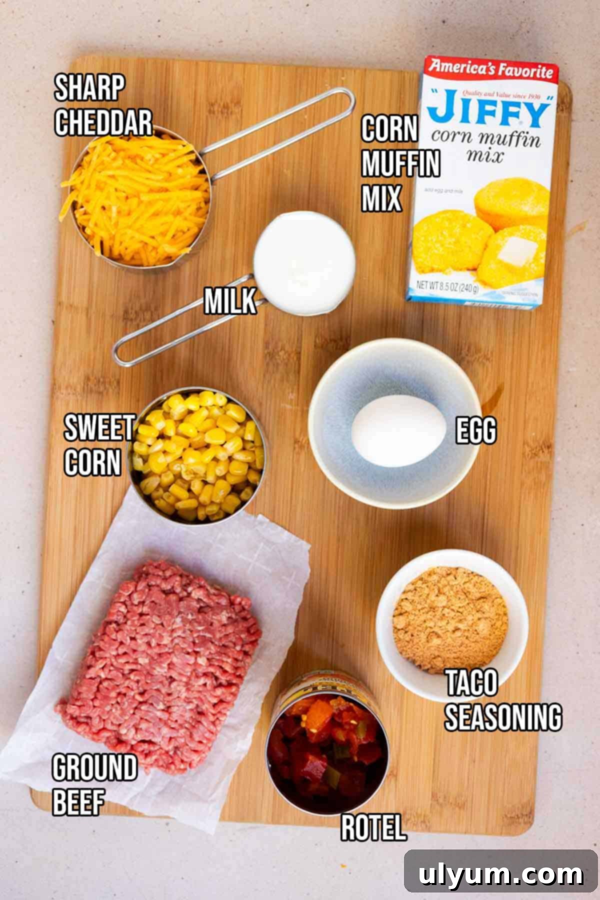All the ingredients laid out for Mexican cornbread casserole: ground beef, Rotel, taco seasoning, Jiffy Corn Muffin Mix, milk, egg, sweet corn, and shredded cheddar cheese.