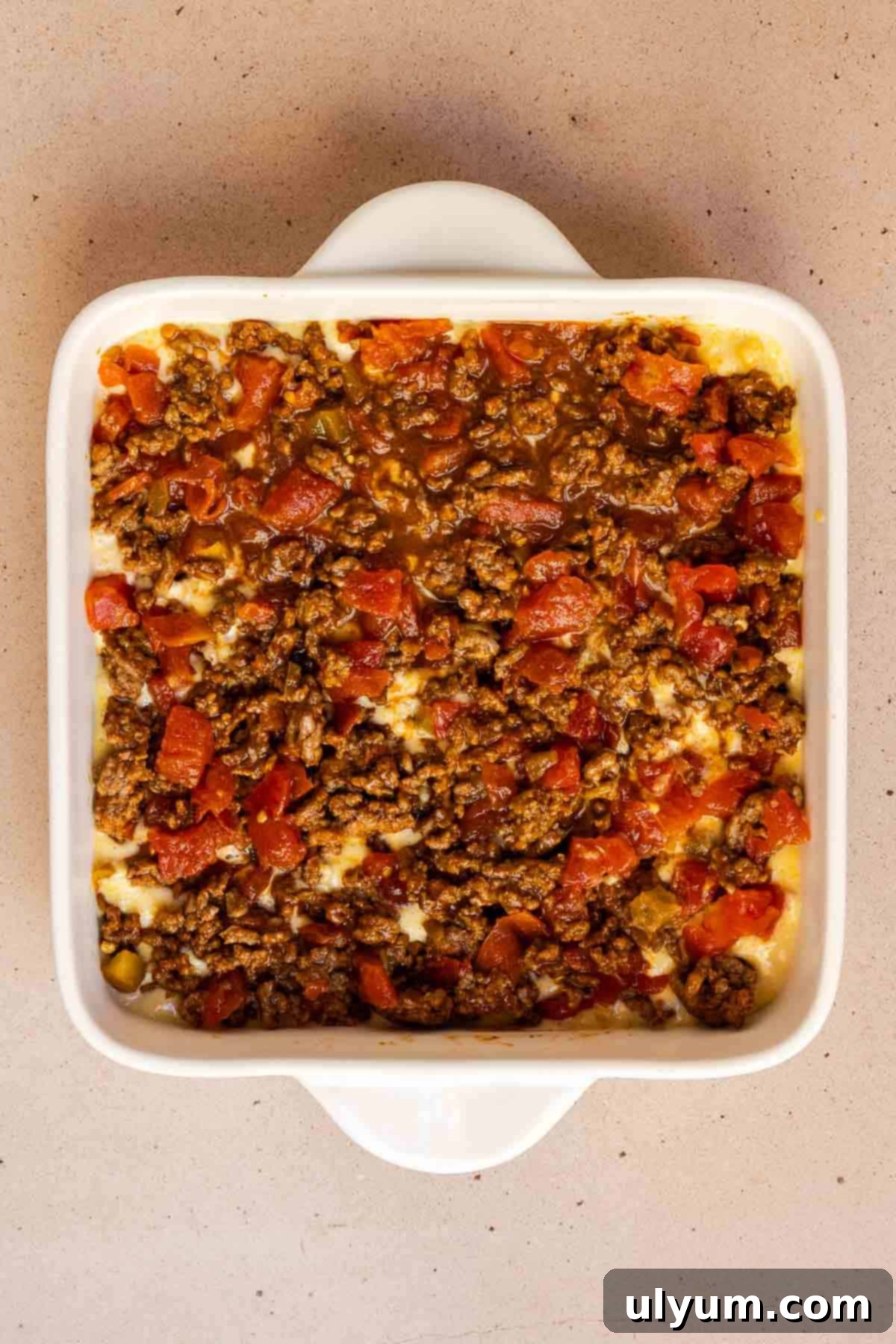 The seasoned ground beef and Rotel mixture is spread over the cornbread base, followed by a layer of shredded cheddar cheese, creating a multi-layered casserole before baking.