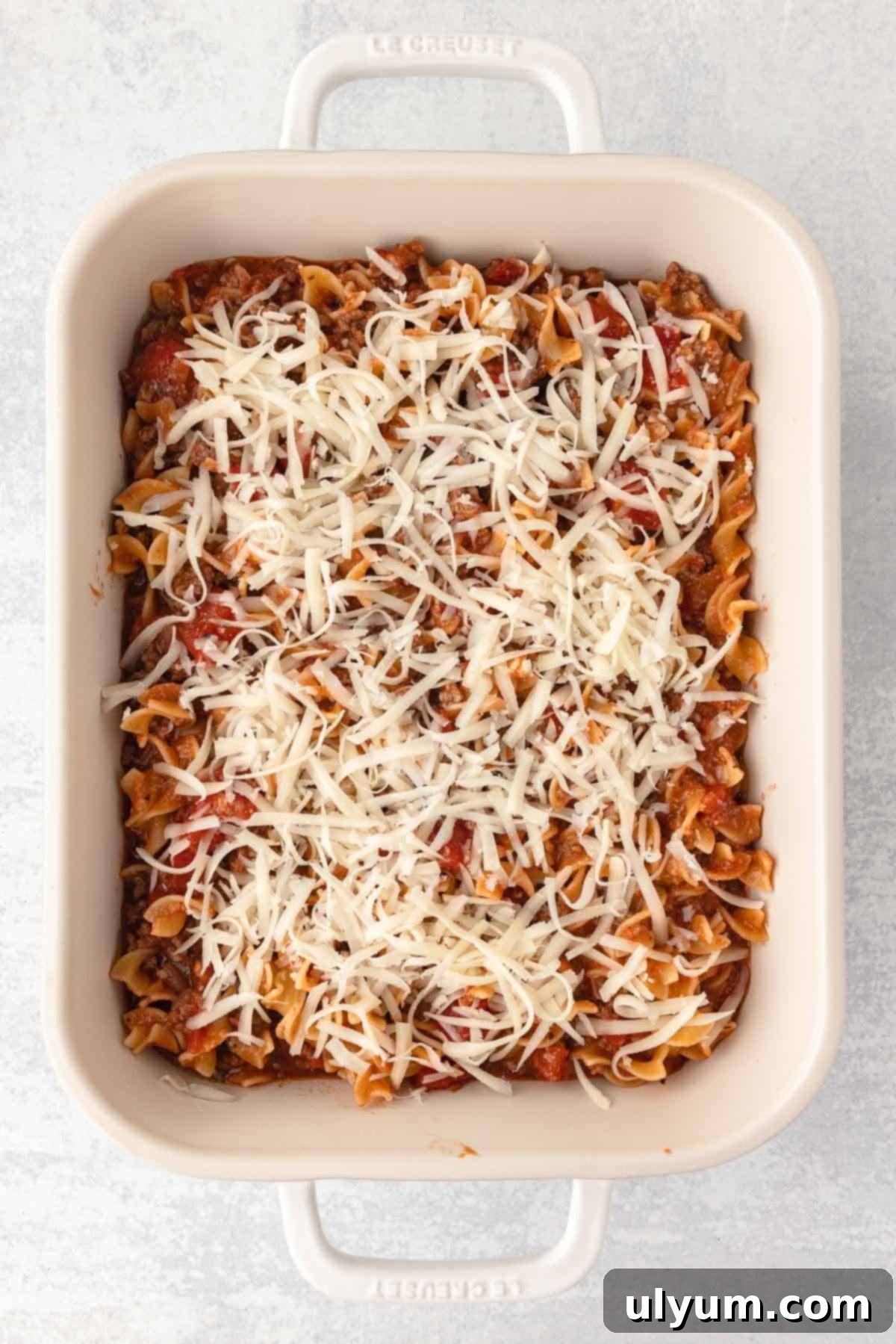 Beef and noodle mixture is spread into a prepared casserole dish and topped with an even layer of mozzarella shreds, ready for baking.