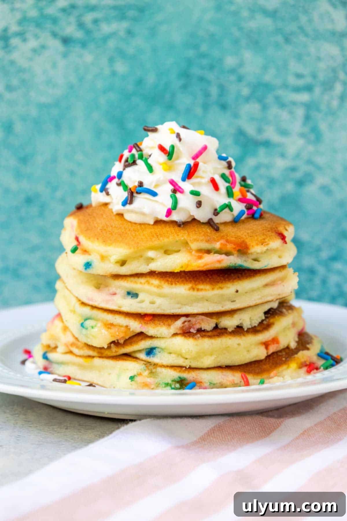 Stack of fluffy Funfetti pancakes topped with whipped cream and rainbow sprinkles, ready to be served for a festive breakfast.