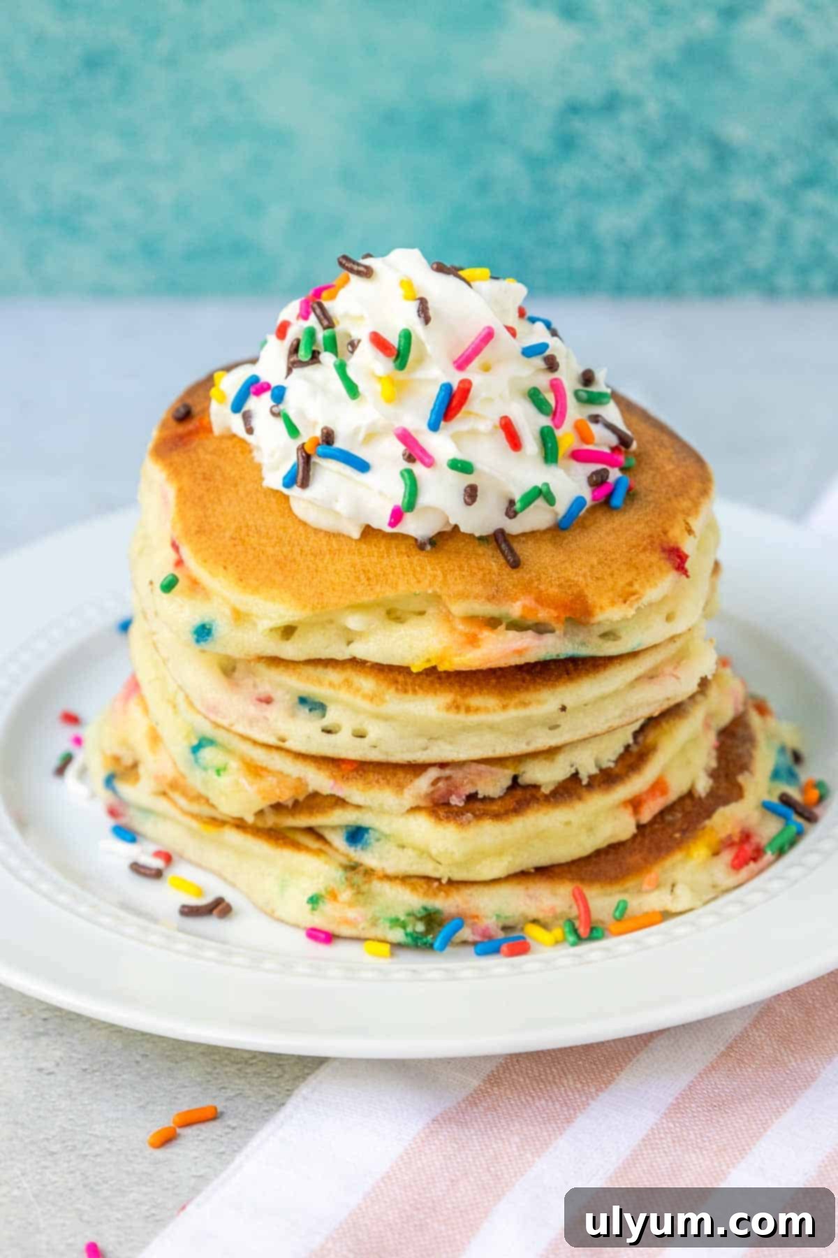 A stack of golden Funfetti pancakes generously topped with fluffy whipped cream and a cascade of rainbow sprinkles.