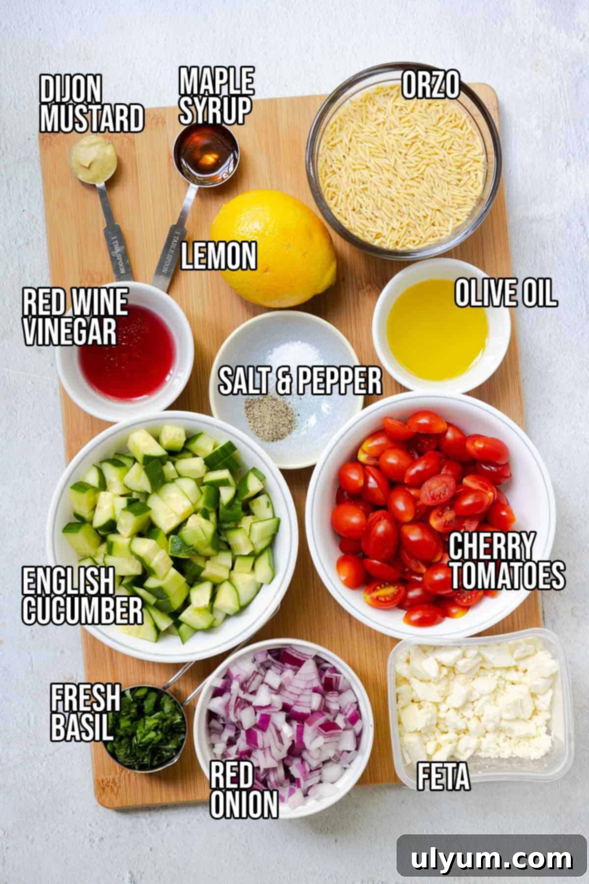 A vibrant display of fresh ingredients for Lemon Feta Orzo Salad: uncooked orzo pasta, whole lemons with zest, a bottle of extra virgin olive oil, red wine vinegar, pure maple syrup, Dijon mustard, salt and pepper shakers, halved bright red cherry tomatoes, neatly diced English cucumber, finely chopped red onion, a block of creamy feta cheese with crumbles, and a bunch of fragrant fresh basil leaves.