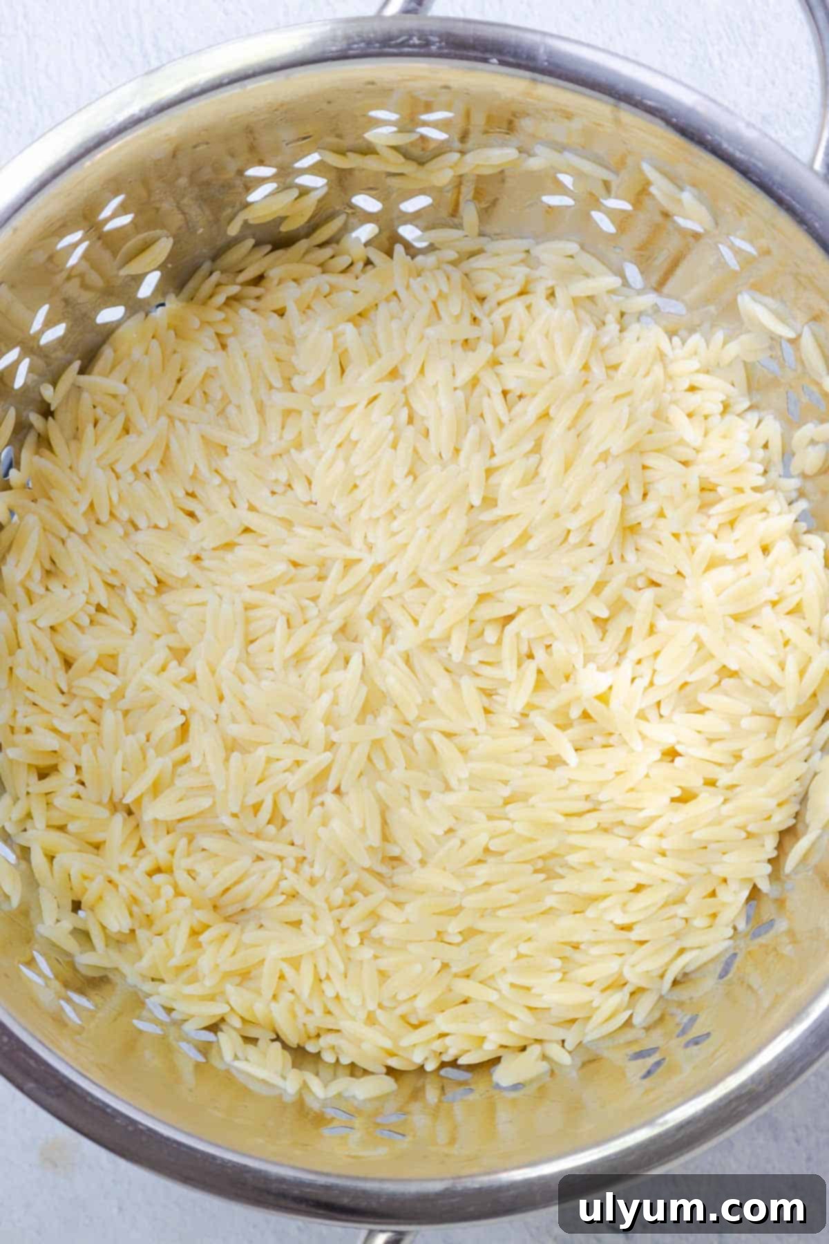 Cooked orzo pasta being drained and rinsed under cold water in a metal colander, steam rising slightly.