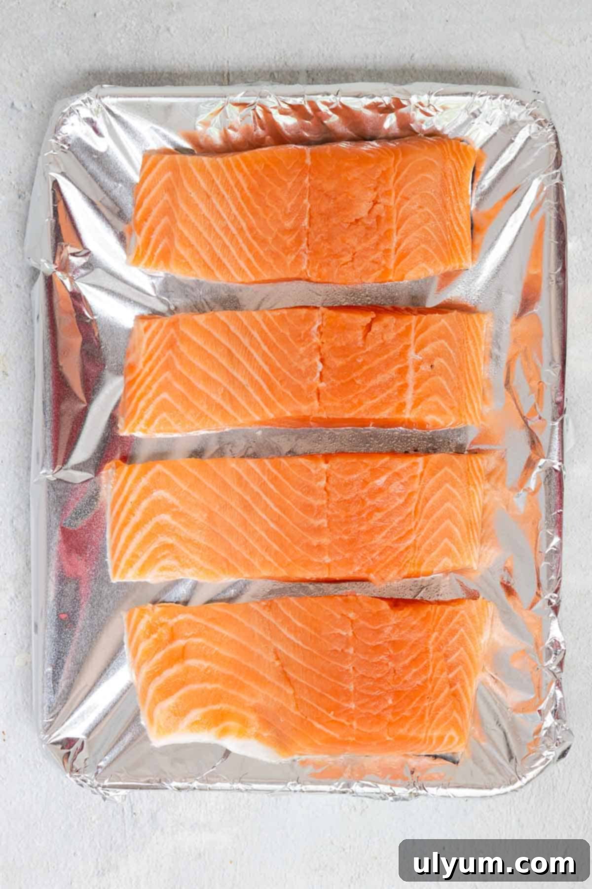 Four raw salmon fillets spaced evenly apart on a baking tray covered with foil and sprayed with cooking spray.