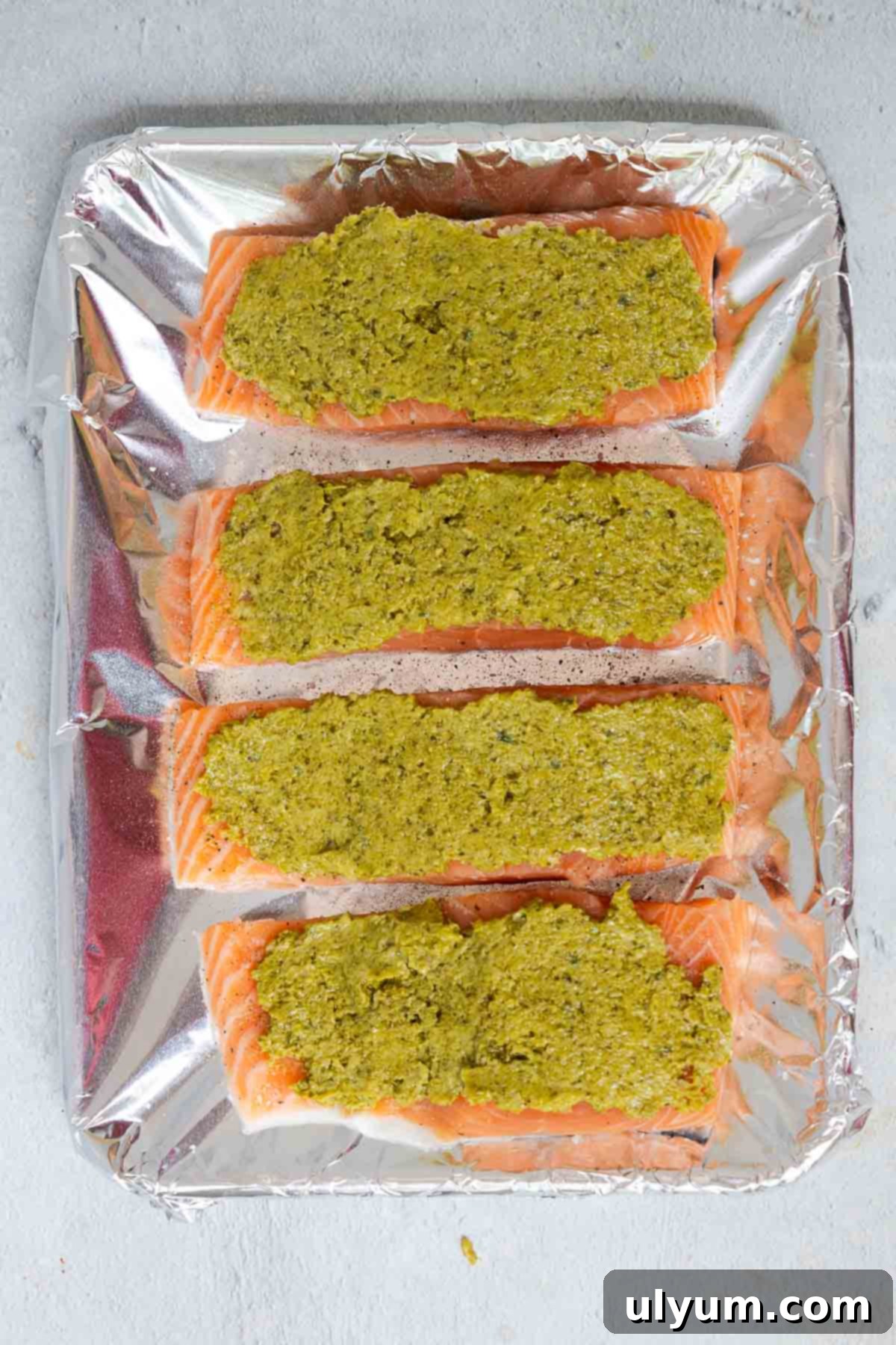 The pesto mixture is spread evenly over top of the uncooked salmon fillets which are evenly spaced on a rimmed baking sheet lined with foil.