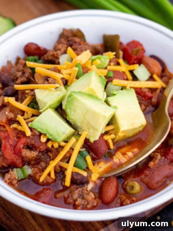 Small bowl of ground beef and sausage chili with shredded cheddar and diced avocado on top.