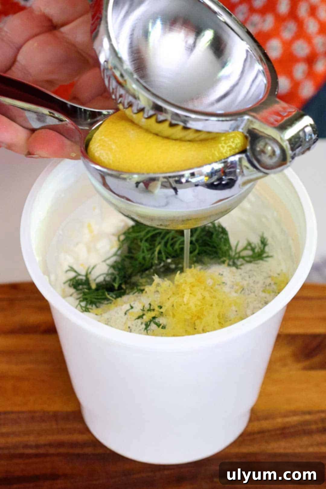 A hand juicing a fresh lemon directly into a container of cottage cheese, which also contains ranch dip mix and fresh dill, illustrating the first step of blending the ingredients for a homemade healthy ranch dip.
