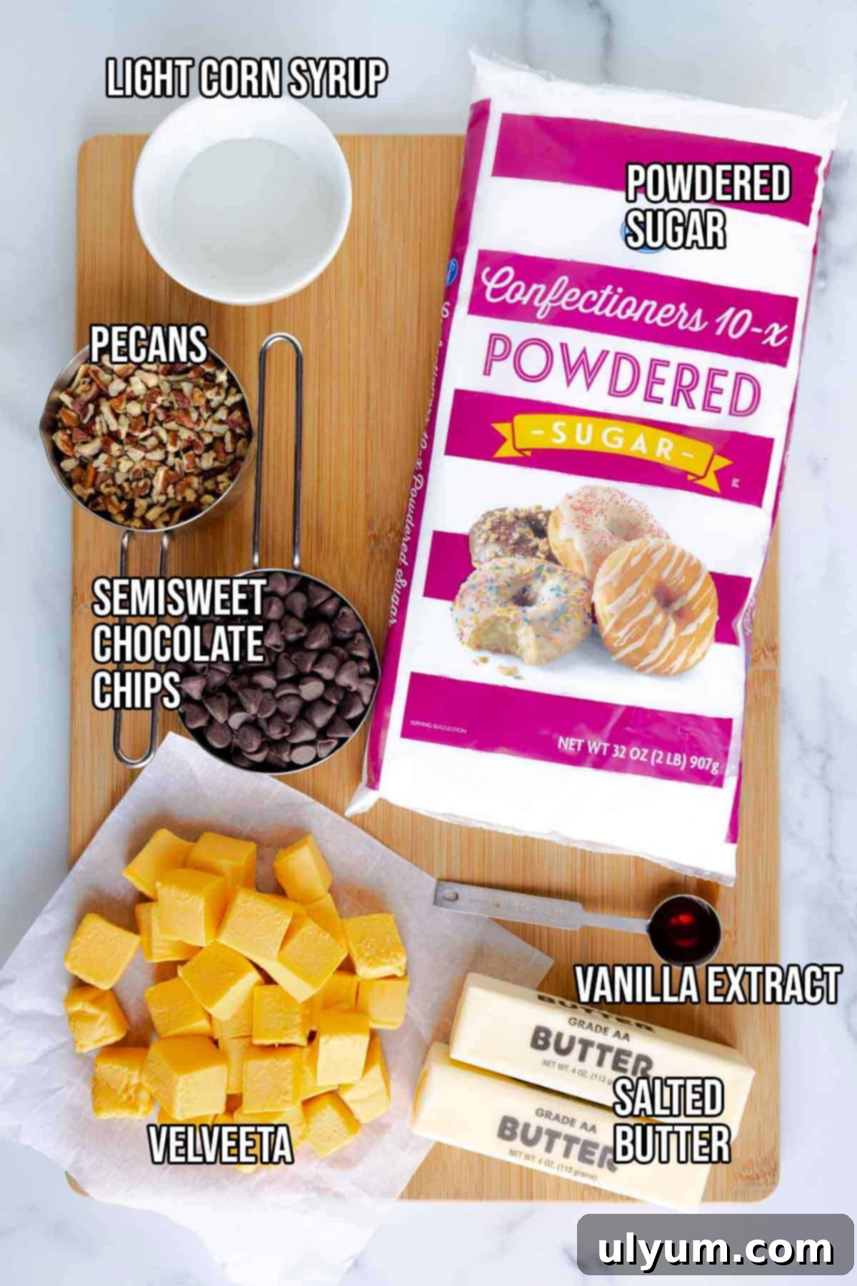 A flat lay photograph of all ingredients for Velveeta fudge: a block of Velveeta, a stick of salted butter, a bowl of semisweet chocolate chips, a small pitcher of light corn syrup, a large bowl of powdered sugar, a bowl of chopped pecans, and a bottle of pure vanilla extract.