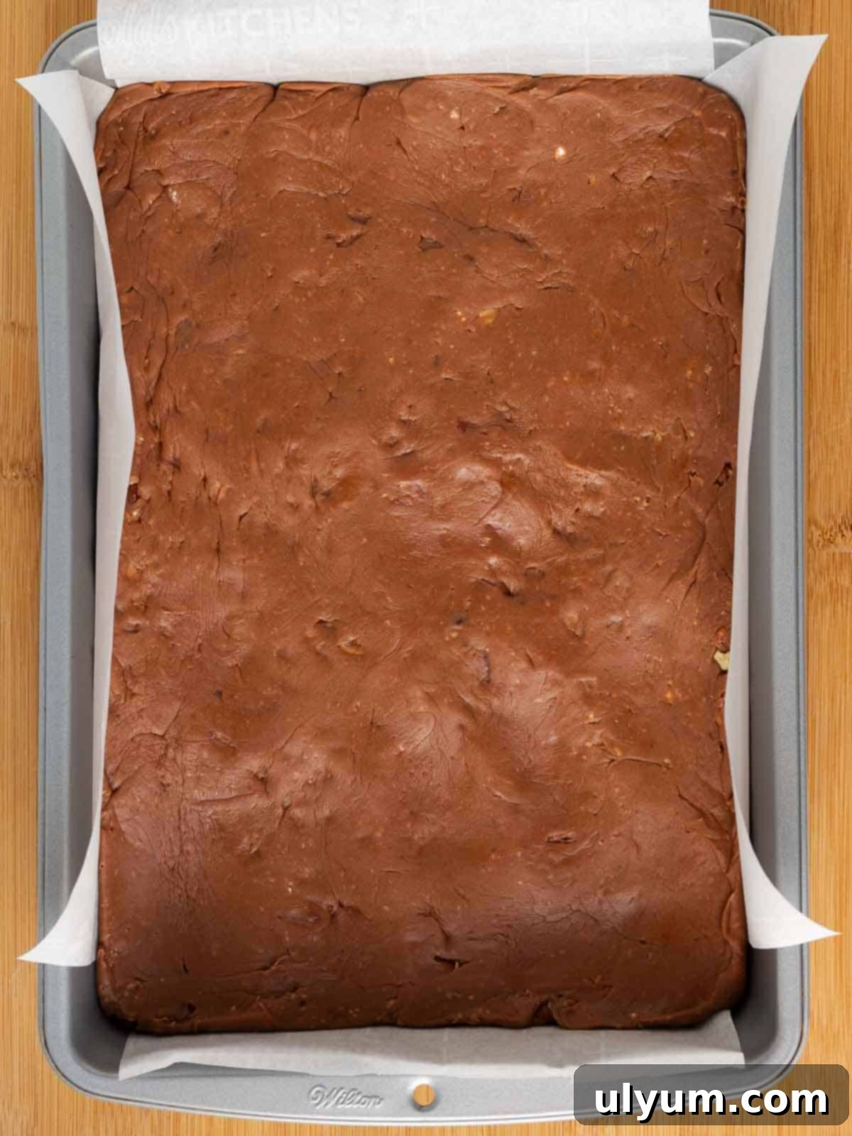 The still-warm chocolate fudge mixture is being poured and spread into a 9 by 13-inch baking pan, which is already lined with parchment paper, for chilling.