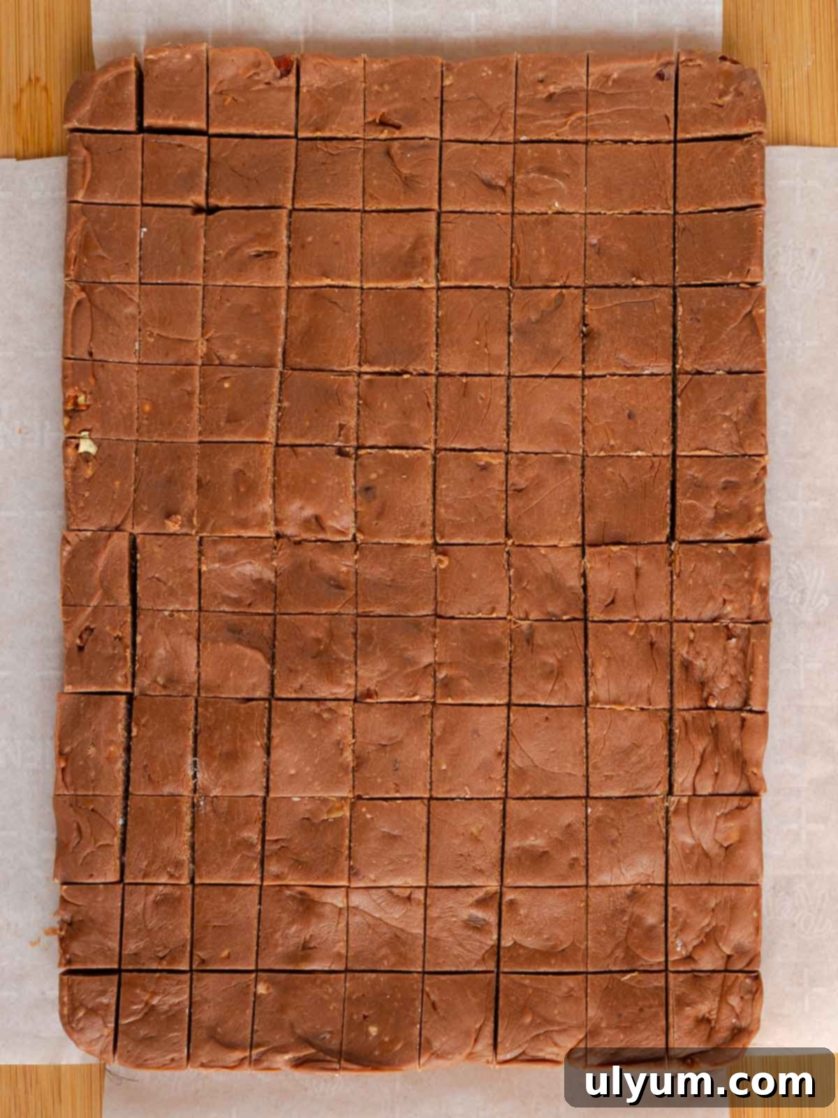 A large slab of firm Velveeta fudge, removed from the baking pan using its parchment paper lining, sits on a cutting board, ready to be cut into 1-inch squares.