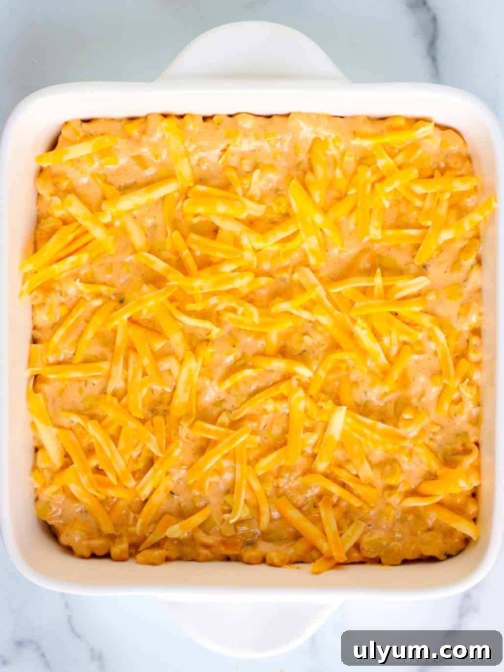Cheesy corn casserole mixture is transferred into in an 8 by 8 inch baking dish and topped with shredded cheese.