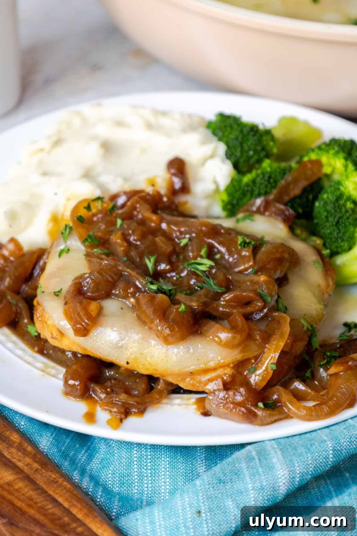 Savory French Onion Pork Chops 2 A pork chop on a plate covered in caramelized onions and cheese with mashed potatoes and broccoli.