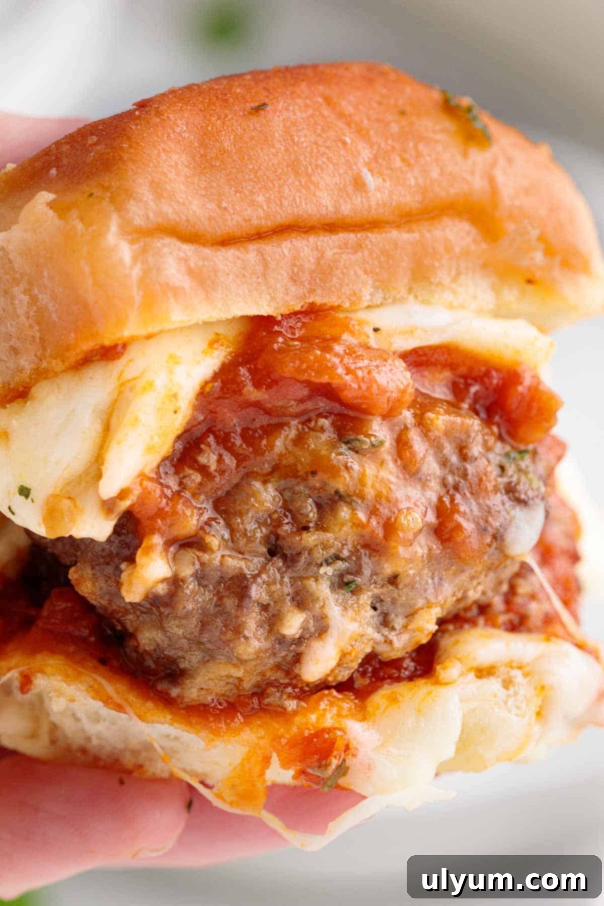 A hand holding a meatball slider with melted cheese and marinara.