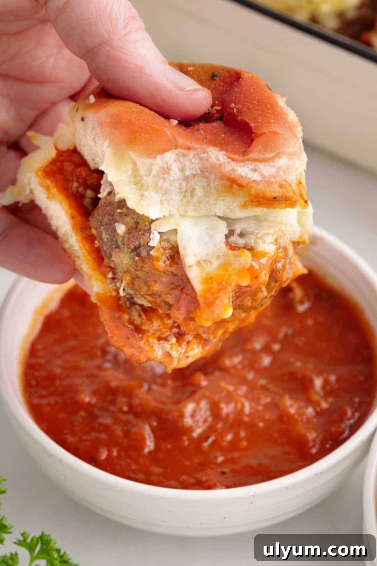 A hand holding a meatball slider that has been dipped into a bowl of marinara sauce.