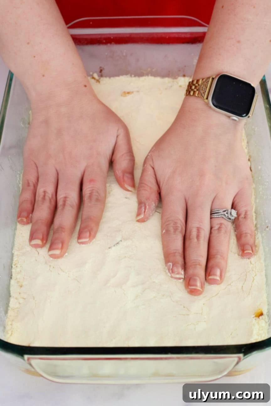 Two hands pressing dry cake mix over the peaches in a baking dish.