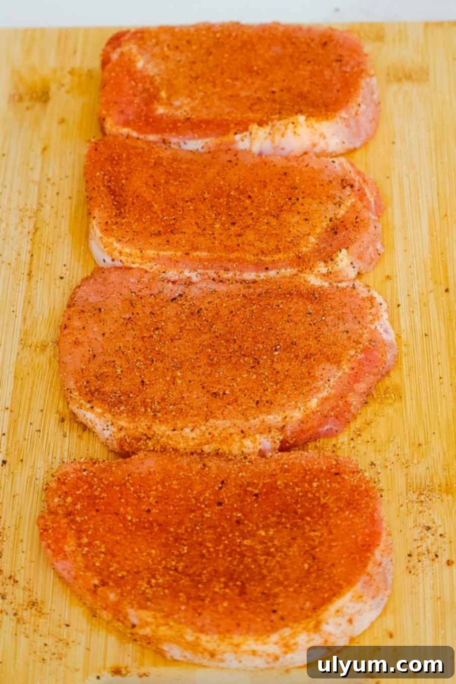 Four raw pork chops seasoned with spices on a cutting board.