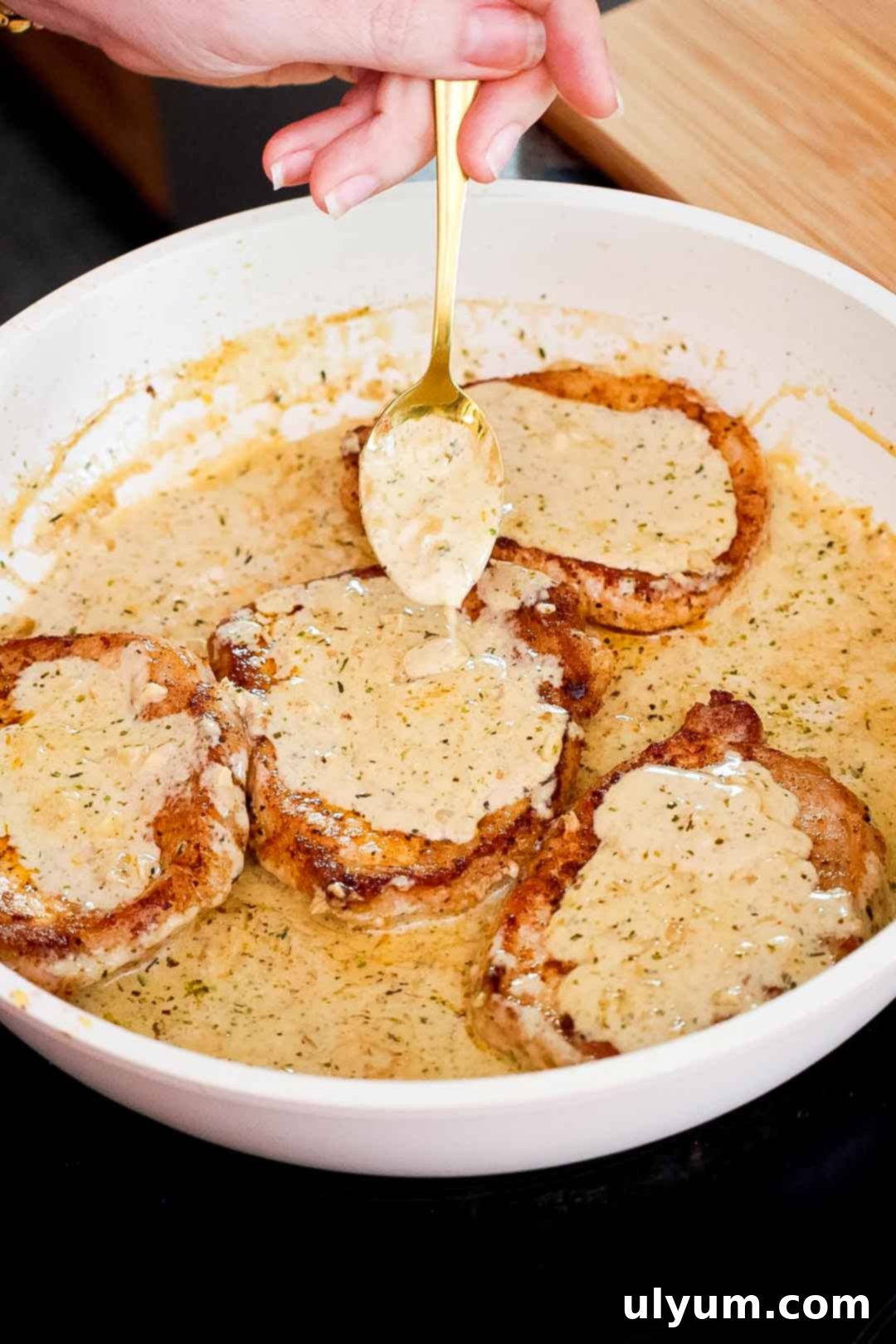 A spoon drizzling garlic cream sauce over pork chops in a skillet.