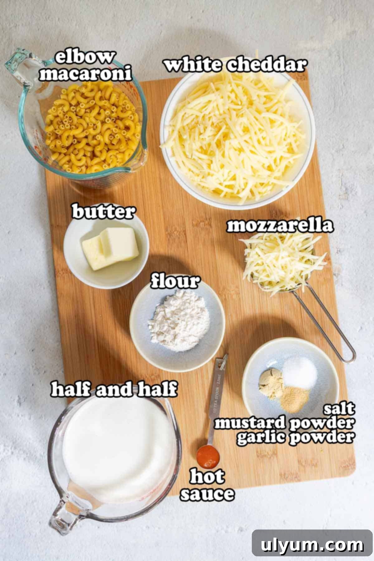 Ingredients for white cheddar mac and cheese laid out on a wooden board, including elbow macaroni, blocks of white cheddar and mozzarella, butter, flour, half and half, hot sauce, salt, mustard powder, and garlic powder.
