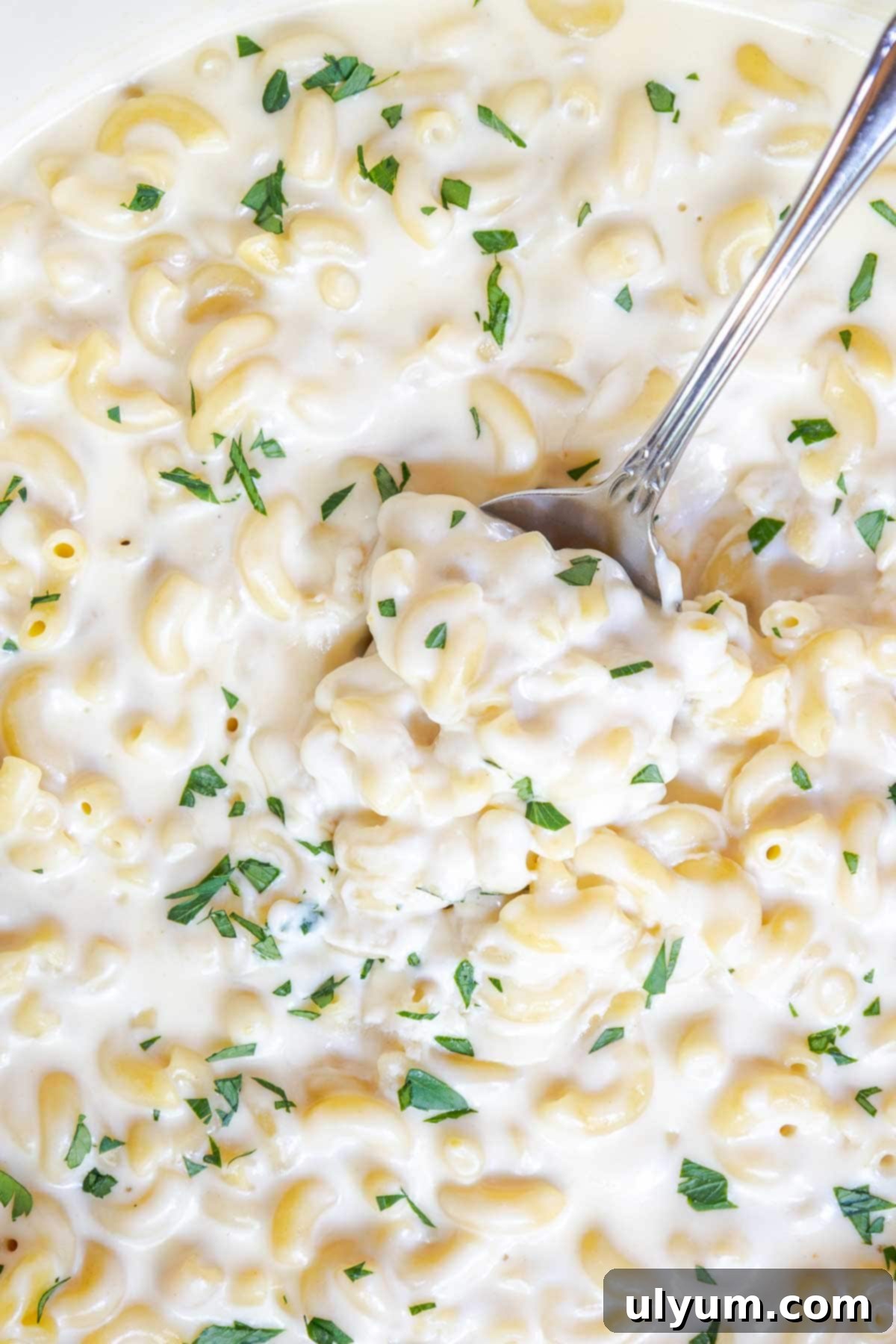 A spoon scooping a generous portion of creamy white cheddar mac and cheese from a pot, garnished with vibrant green parsley, highlighting its rich texture.