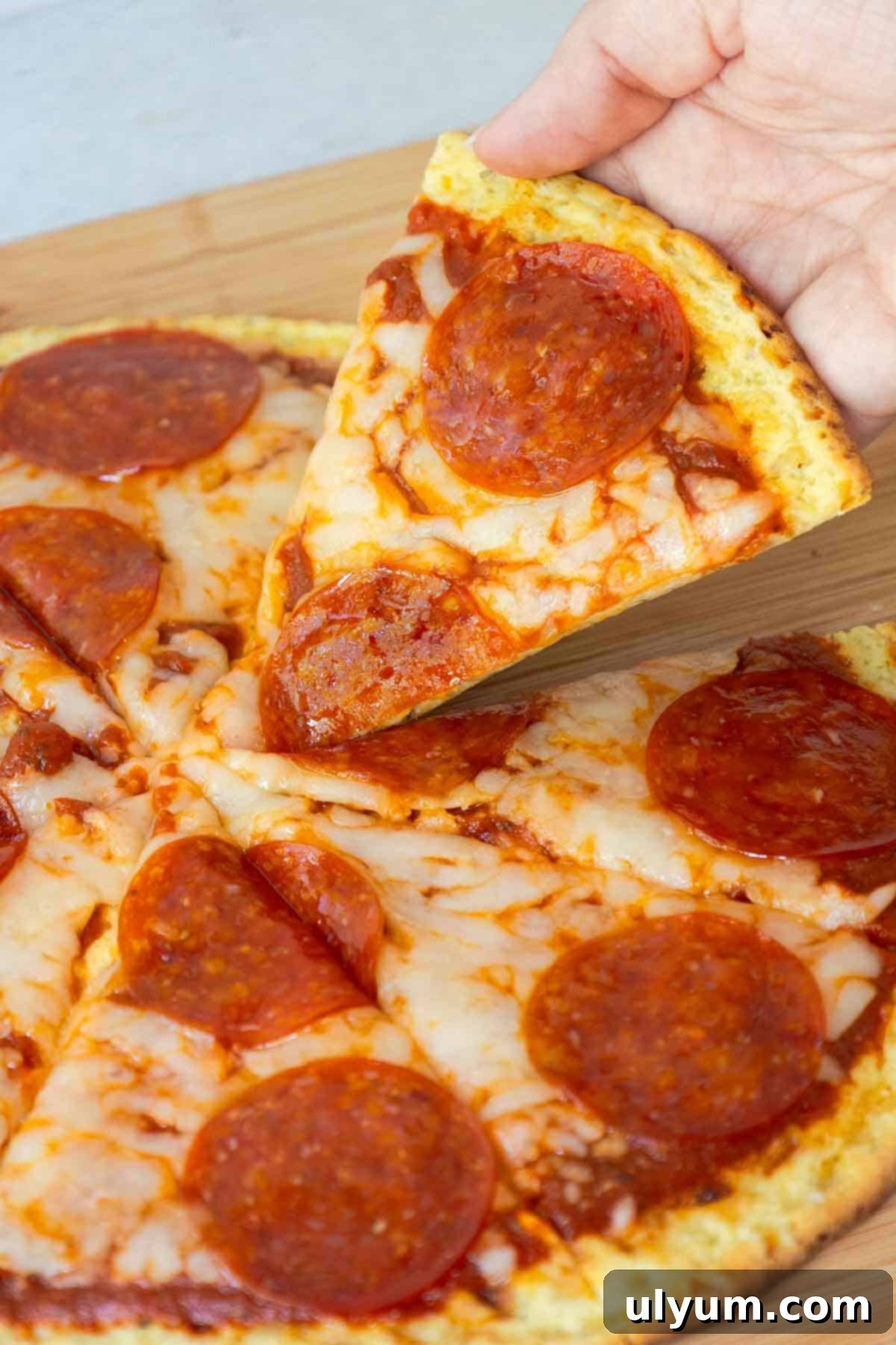A hand lifting a perfect slice of pepperoni pizza from the rest of the pie, showcasing the crispy cottage cheese crust.
