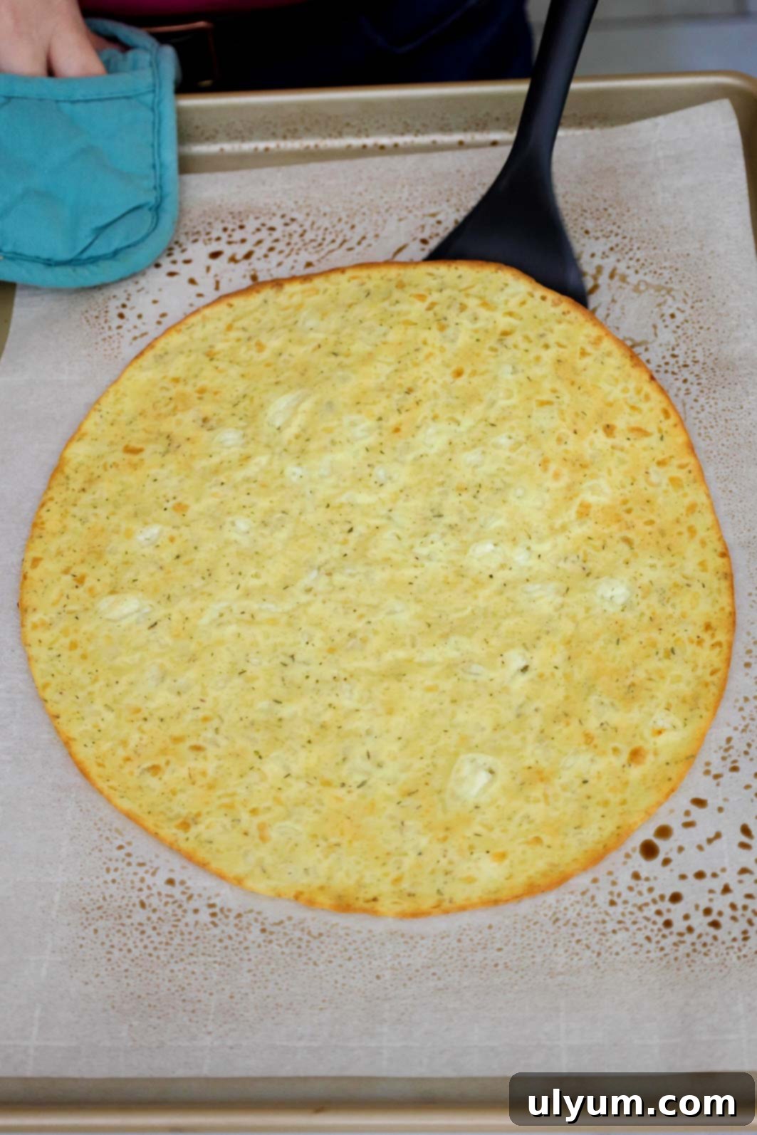 A partially baked pizza crust on parchment paper being flipped with a large spatula to ensure even browning and crispness.