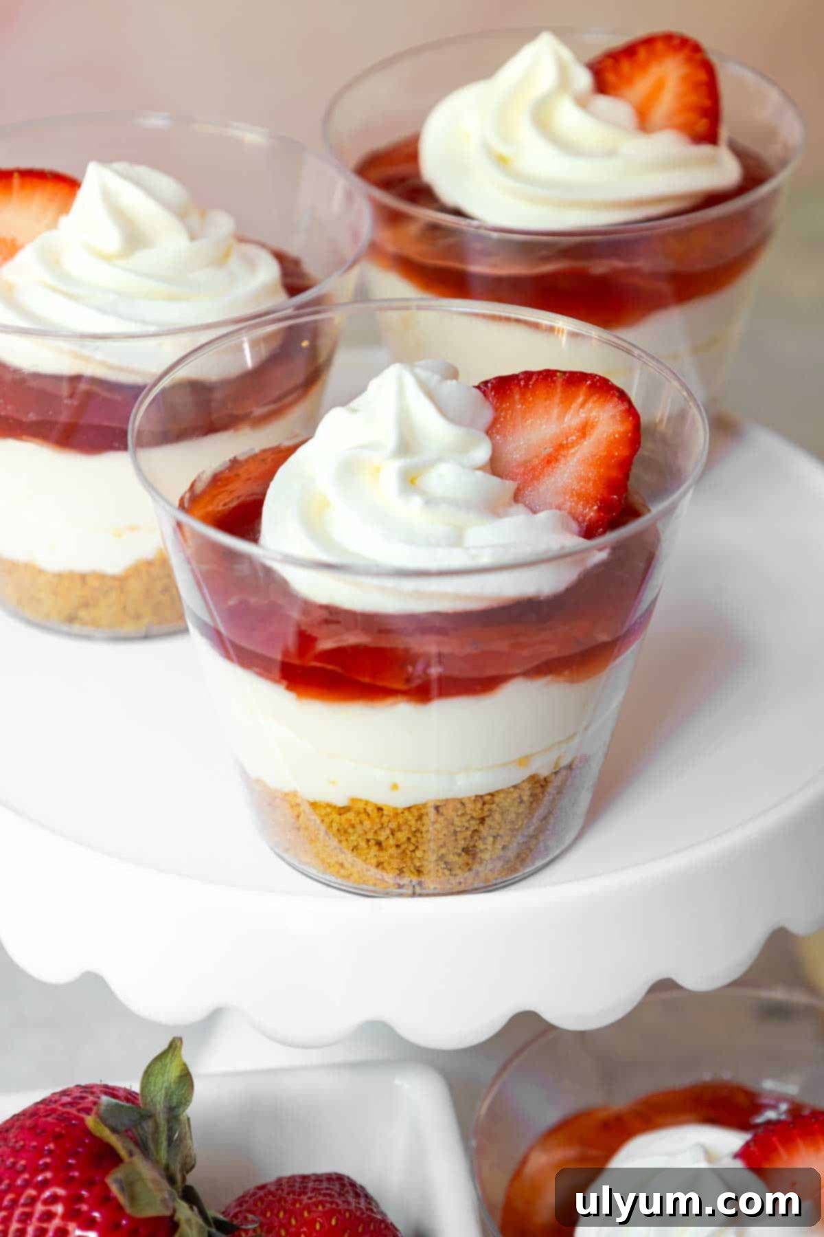 Cheesecake cups with a graham cracker base, creamy filling, strawberry topping, and whipped cream, each topped with a sliced strawberry, perfect for a no-bake summer dessert.
