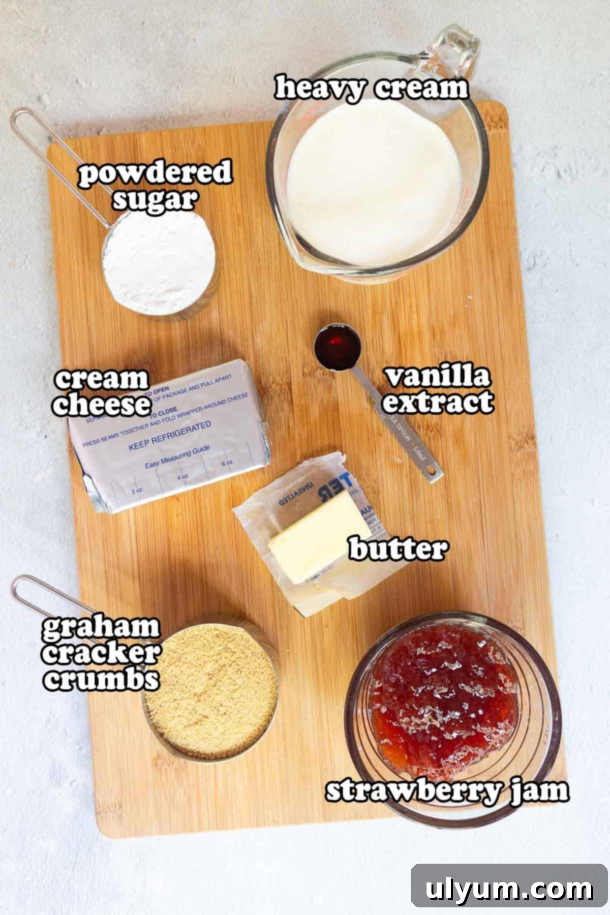 Ingredients for no-bake strawberry cheesecake cups laid out on a wooden board: cream cheese, powdered sugar, heavy cream, vanilla extract, butter, graham cracker crumbs, and strawberry jam.