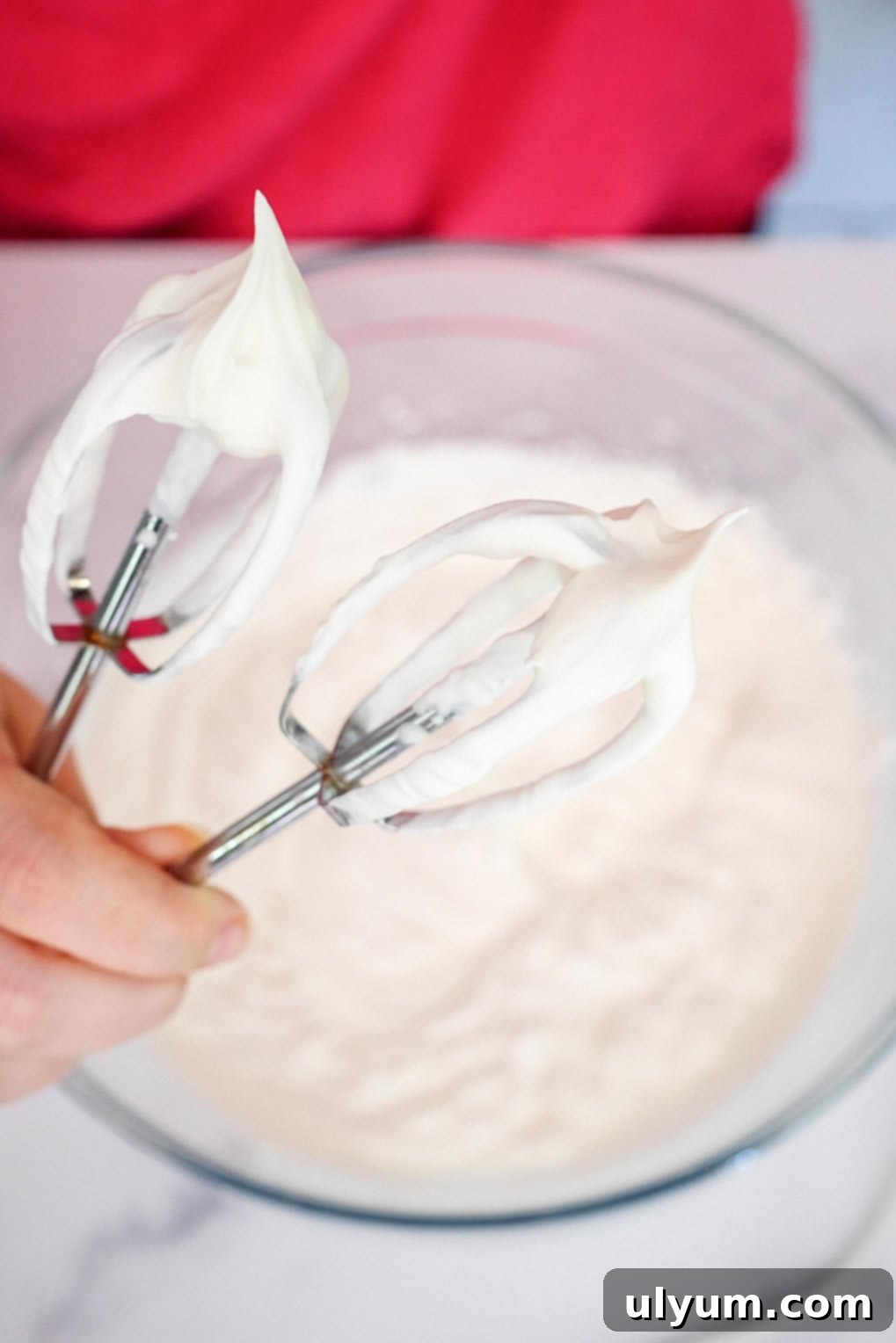 Electric beaters creating stiff whipped cream peaks in a large glass mixing bowl.
