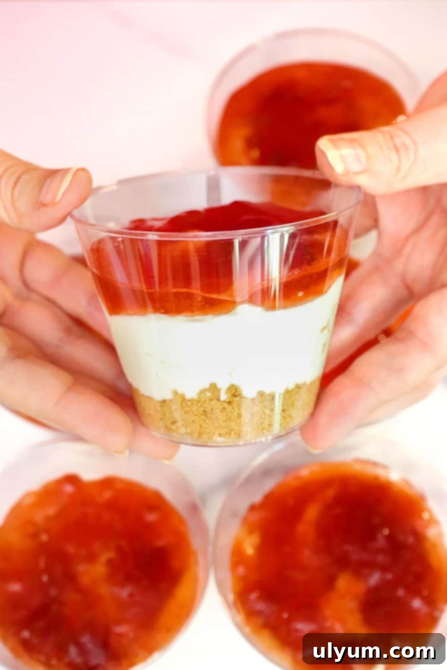 Hands holding a cup layered with graham cracker crust, cheesecake filling, and strawberry jam, demonstrating the assembly process.