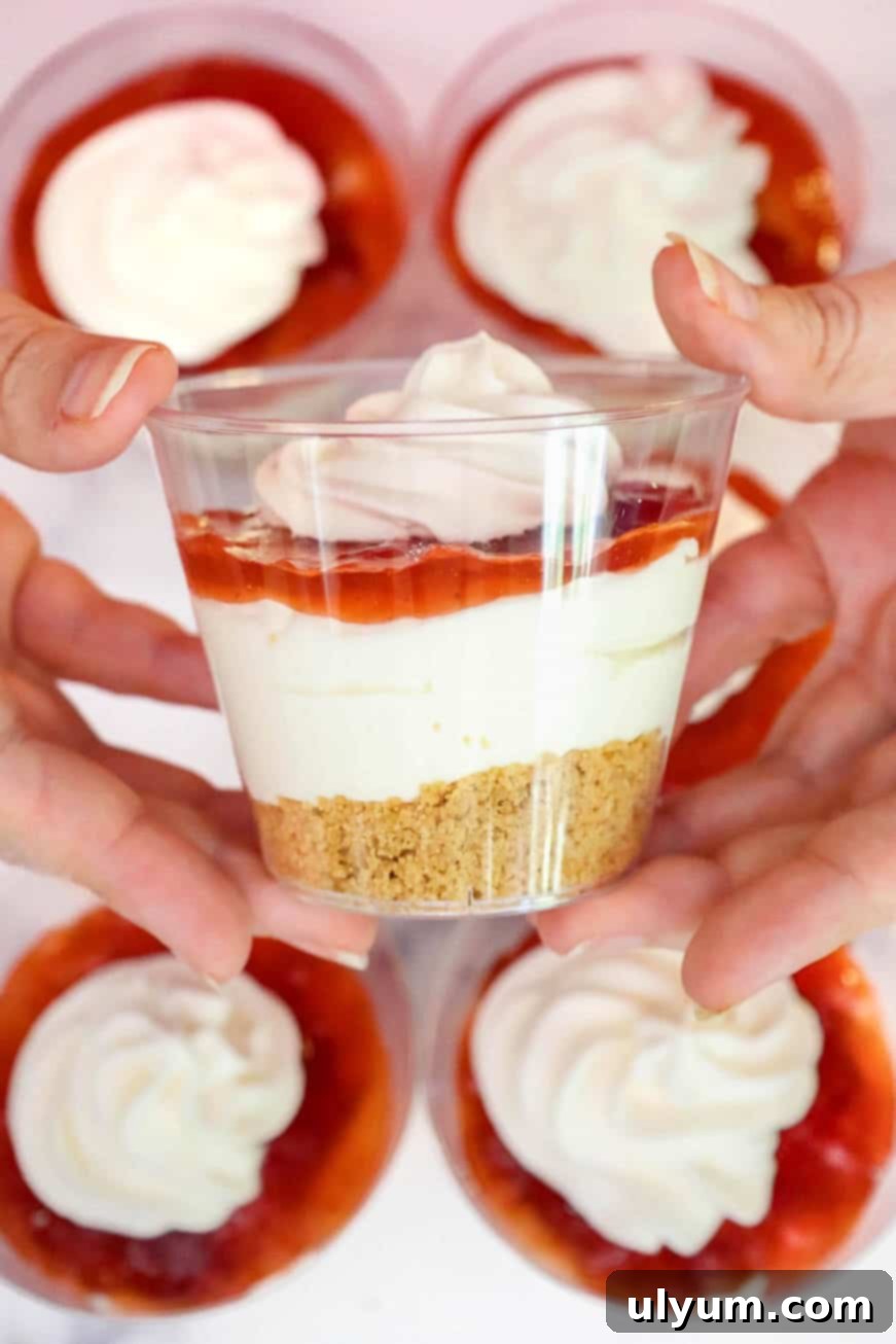 Hands holding a finished strawberry cheesecake cup topped with whipped cream, with other prepared cups in the background.