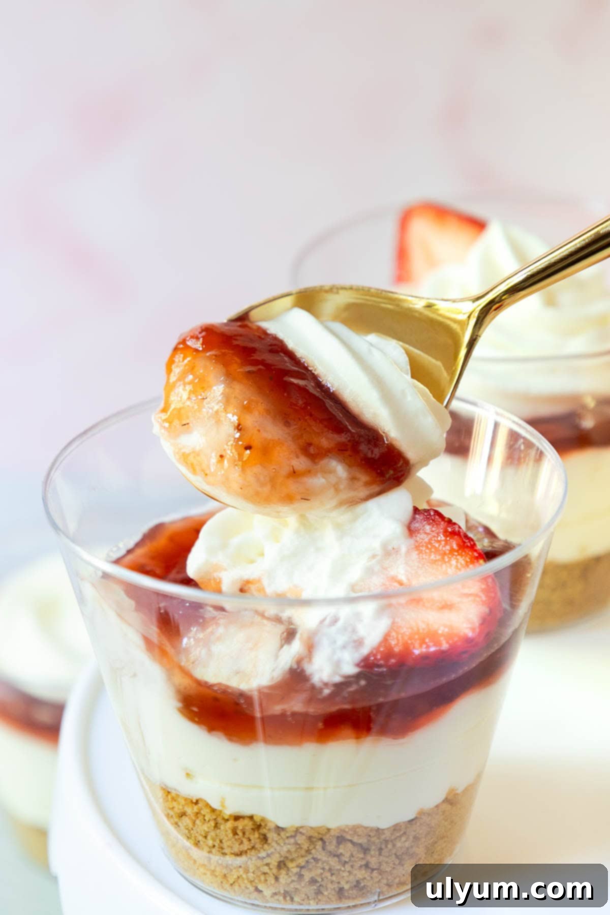 A spoon lifting a perfect bite of strawberry cheesecake from a cup, revealing layers of creamy filling, jam, and a fresh strawberry slice on top.