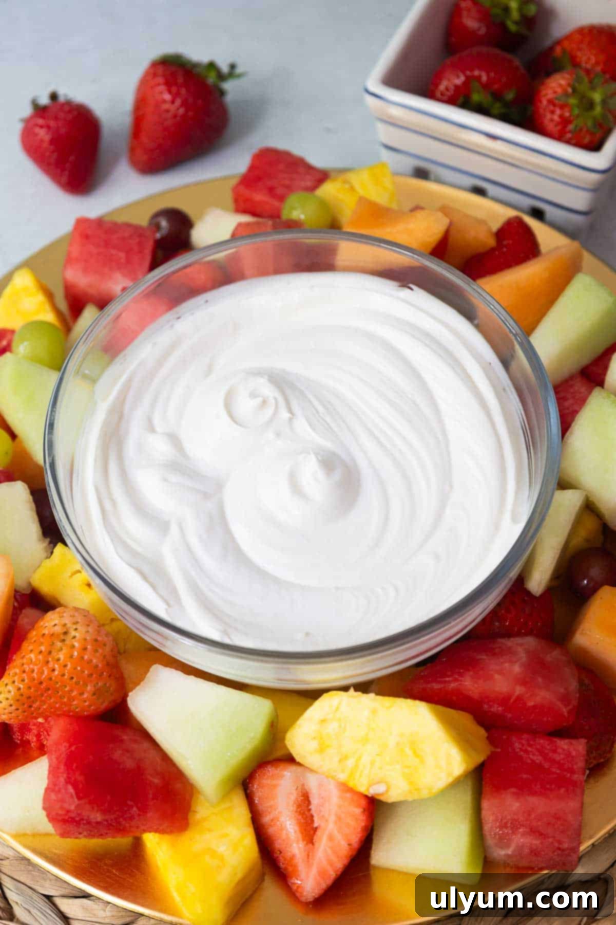 A bowl filled with fluffy white fruit dip, surrounded by an assortment of fresh fruit including strawberries, pineapple, cantaloupe, honeydew, grapes, and watermelon.