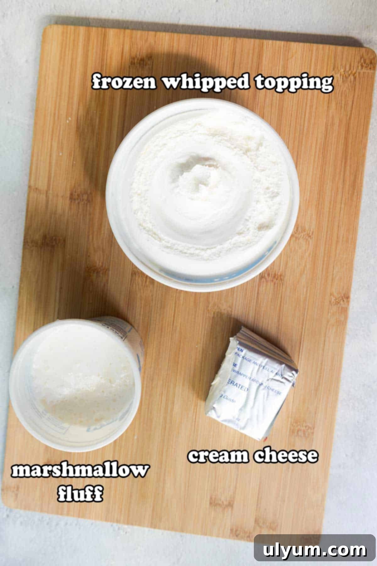 Overhead view of three ingredients labeled for fruit dip: a tub of frozen whipped topping, a jar of marshmallow fluff, and a half block of cream cheese on a wooden cutting board.