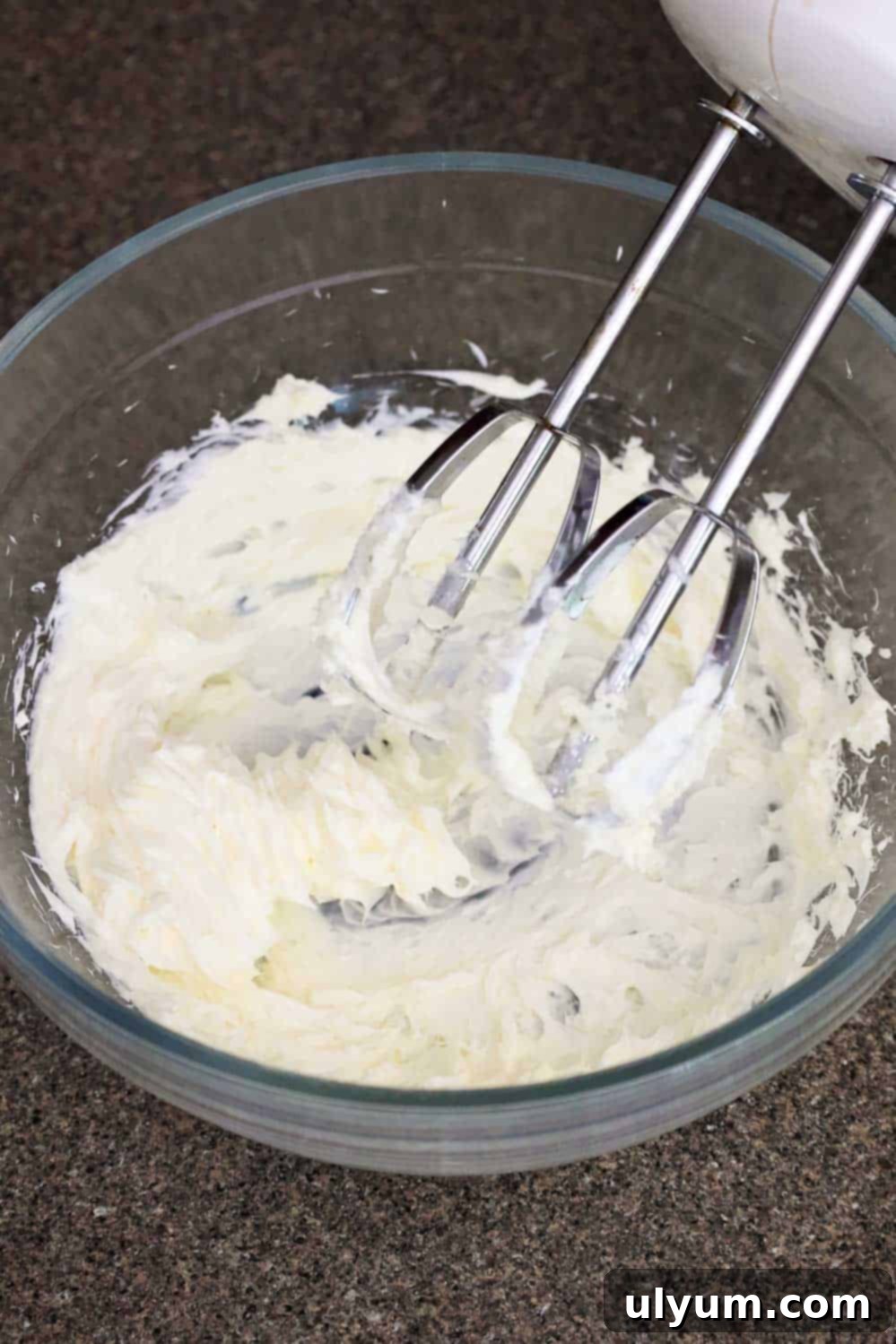 A bowl of cream cheese being mixed with an electric hand mixer.