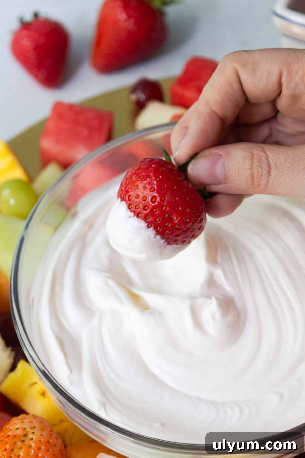 A hand holding a fresh strawberry dipped halfway into a creamy white fruit dip, with a colorful fruit platter visible in the background.